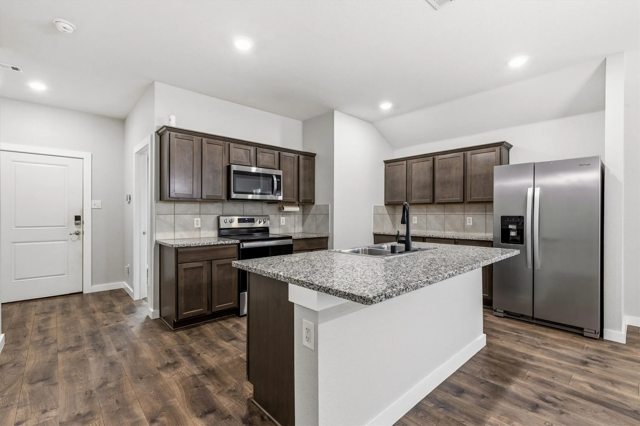 Kitchen featuring wood-finish flooring, dark wood cabinetry, granite countertops, an island with an undermount sink, and stainless steel appliances