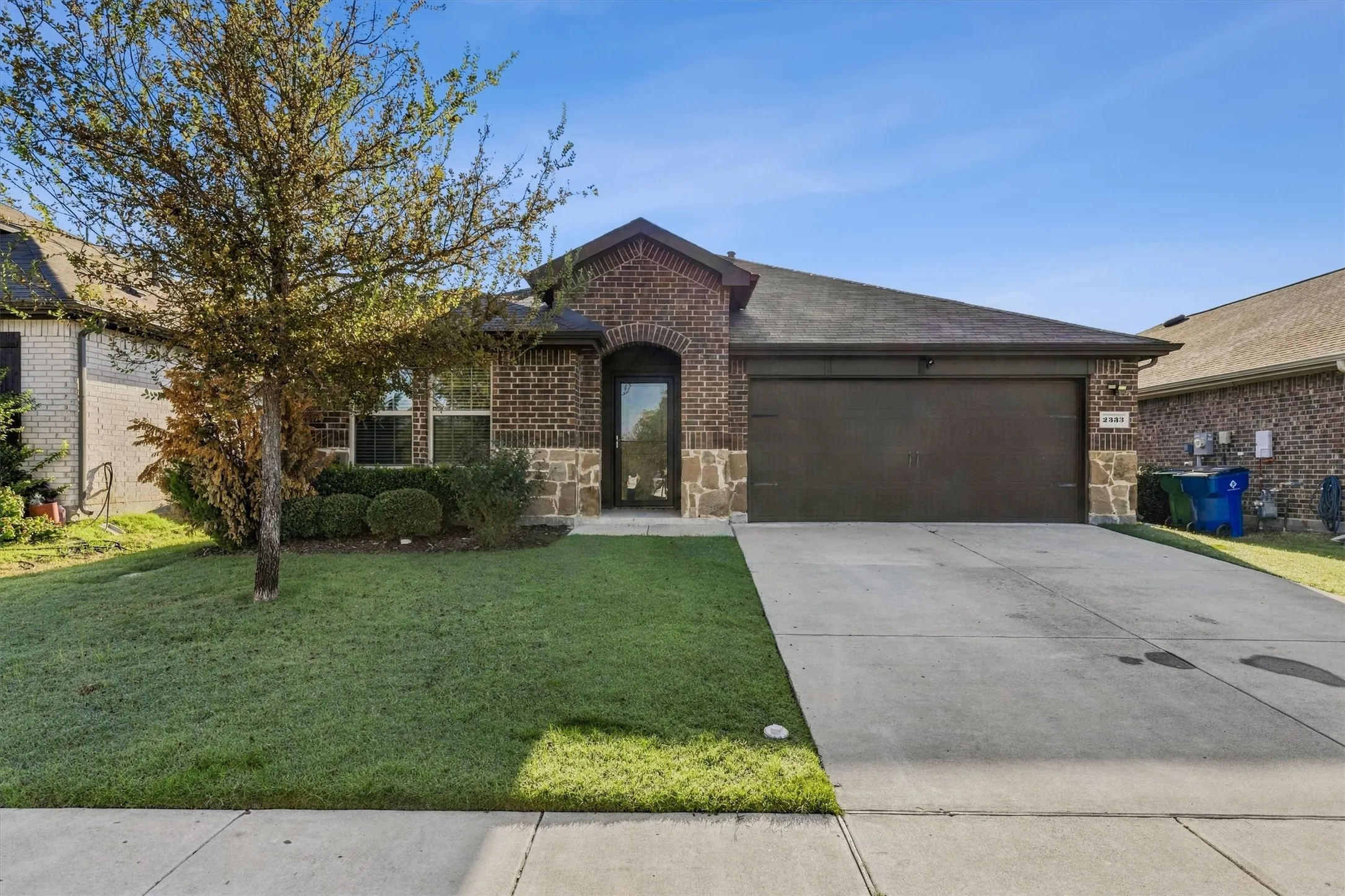 Brick facade with stone accents, arched entry, attached garage, concrete driveway, and front lawn with mature landscaping