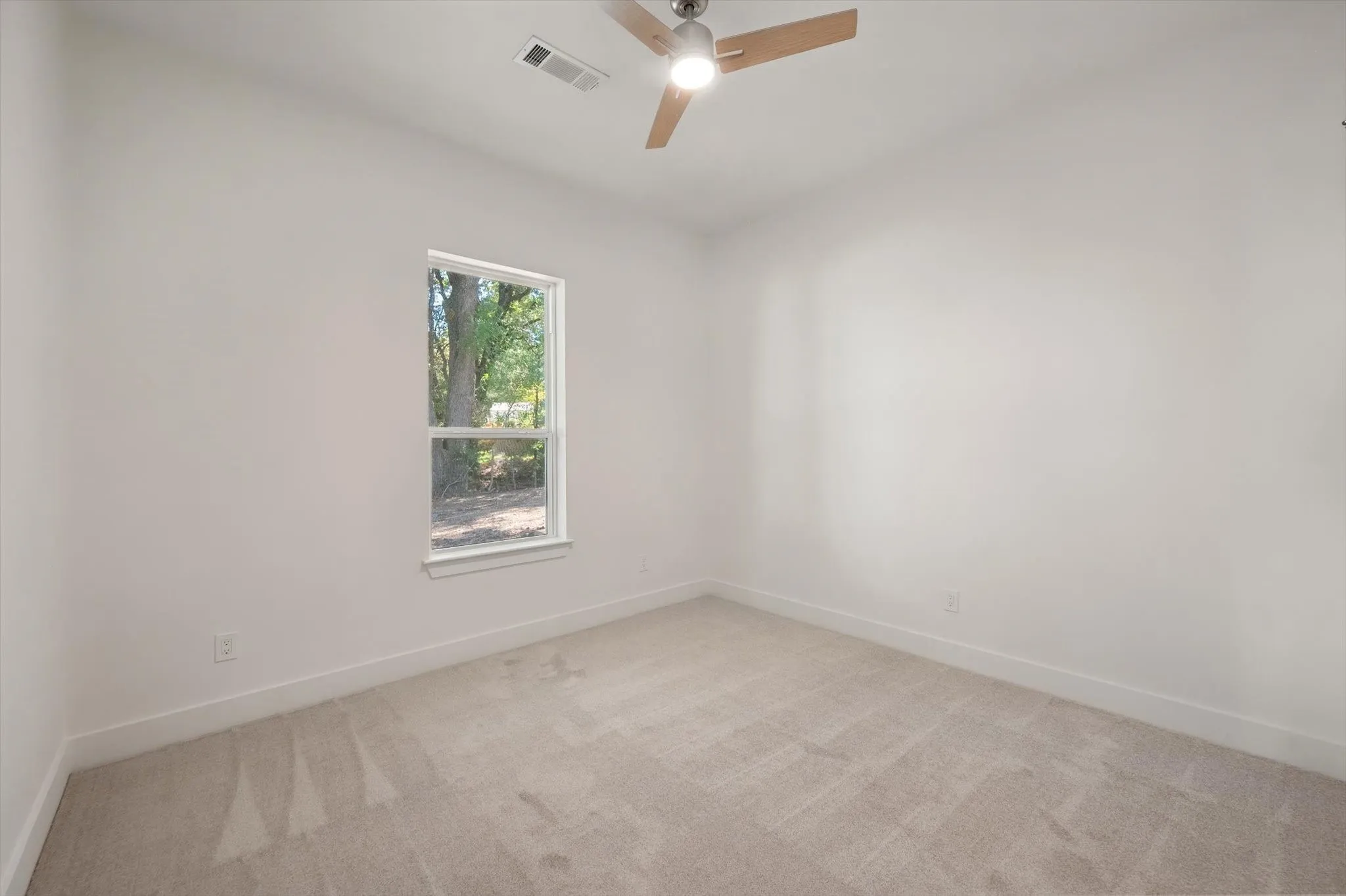 Bright guest bedroom featuring clean white walls, plush carpeting, ceiling fan, and tree-lined views.
