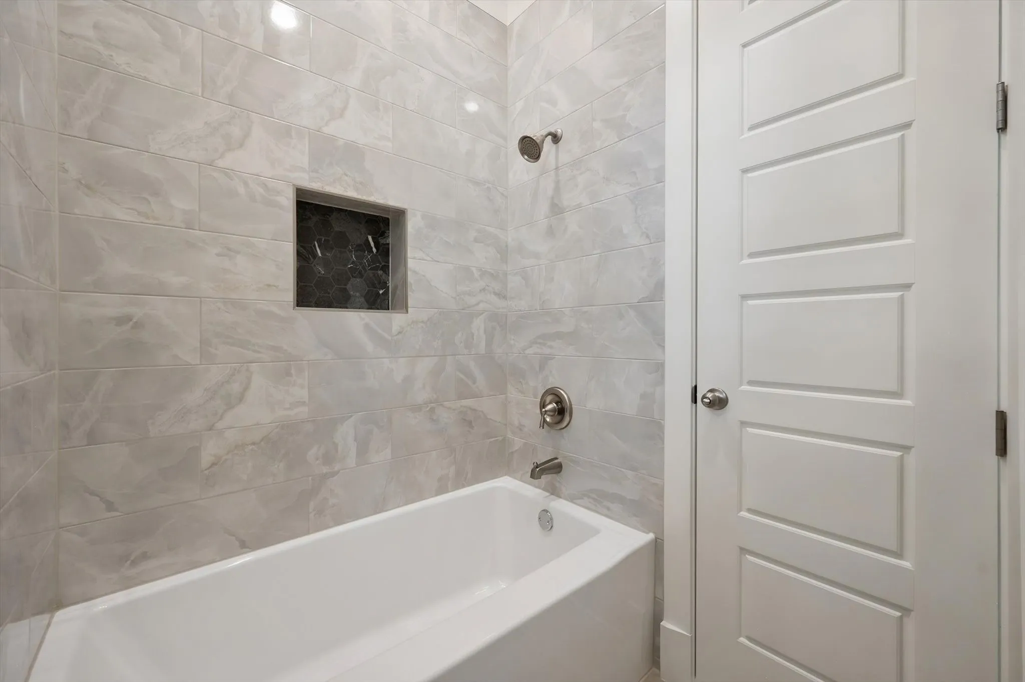 Soaking tub and shower combo surrounded by marble-patterned tile walls and a contrasting black hexagon accent niche.