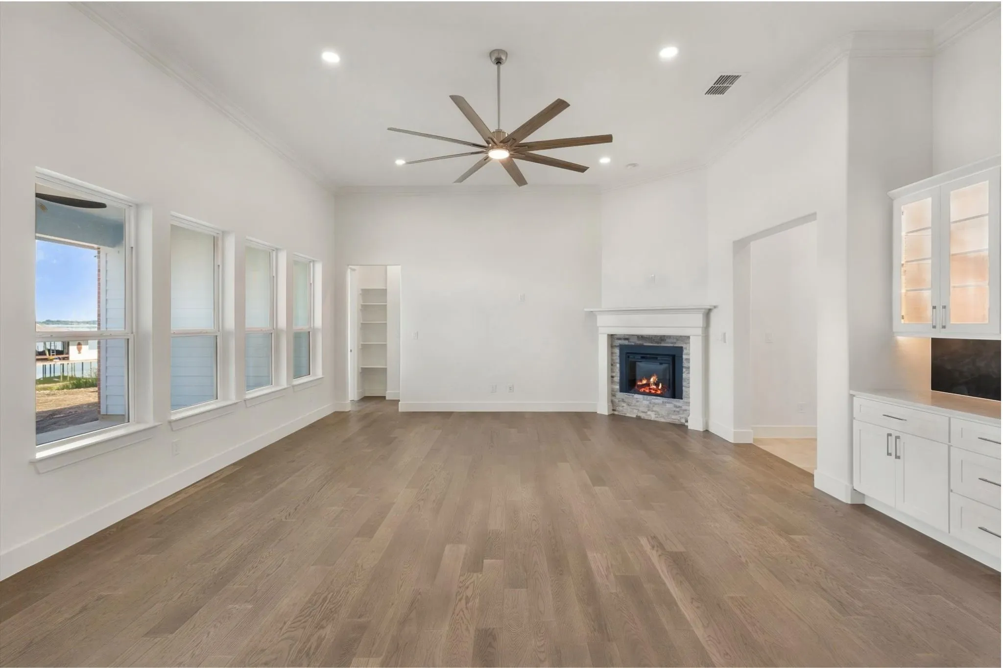Spacious living area highlighted by a modern ceiling fan, warm fireplace glow, and wall-to-wall lake view windows.