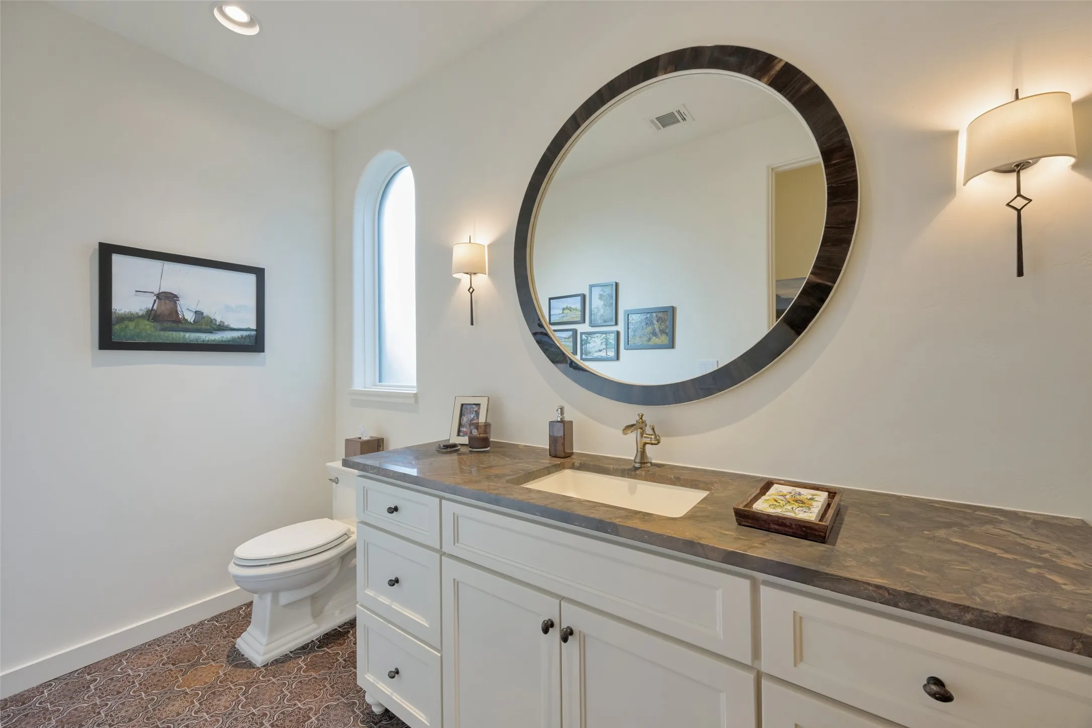 Arched window with casing, large round mirror with dark frame, white vanity with integrated sink, stone-finish countertop, and patterned floor tile