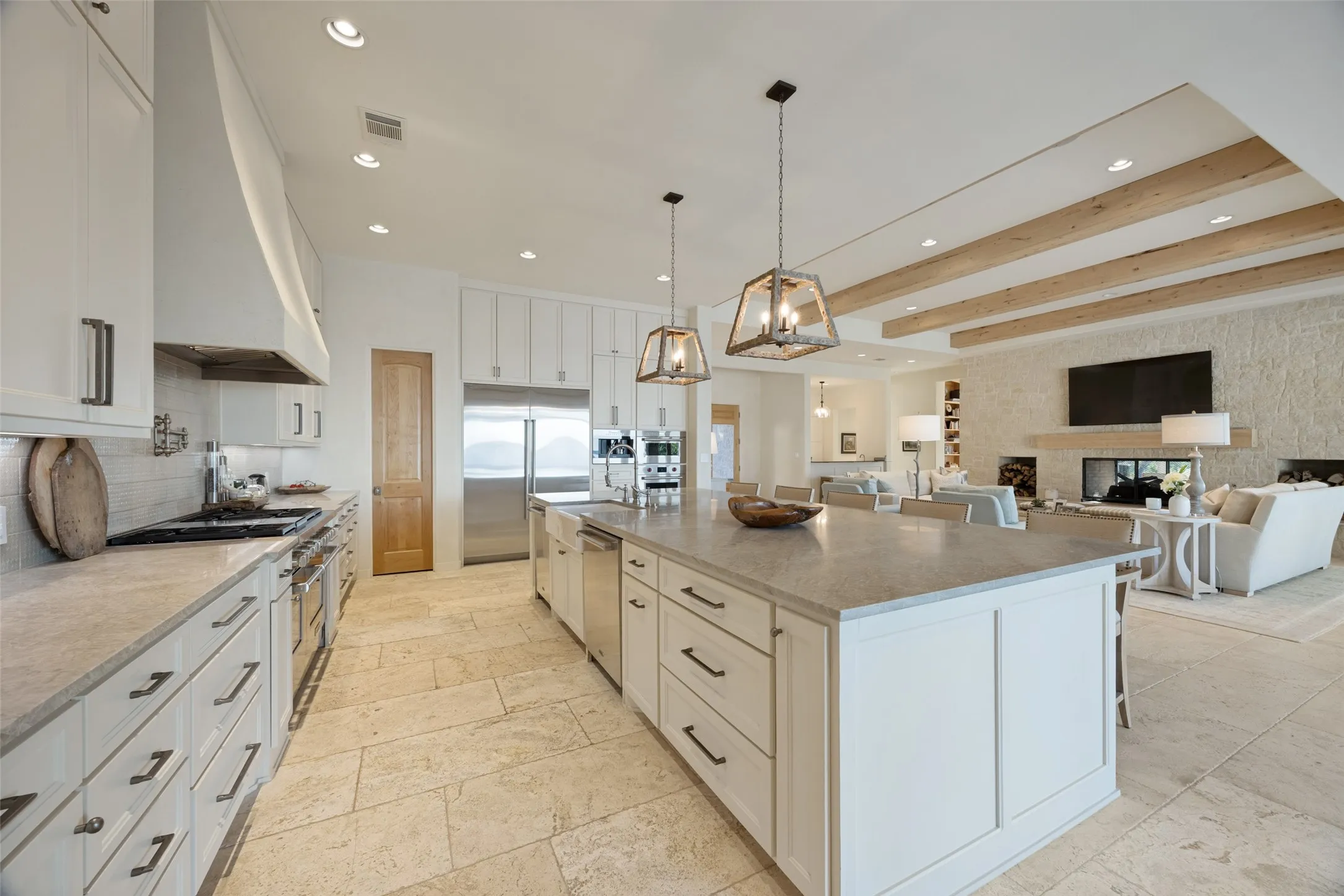 Gourmet kitchen with stainless steel appliances, expansive central island, recessed lighting, and tile flooring, opening to a living area with exposed ceiling beams and a stone-clad fireplace
