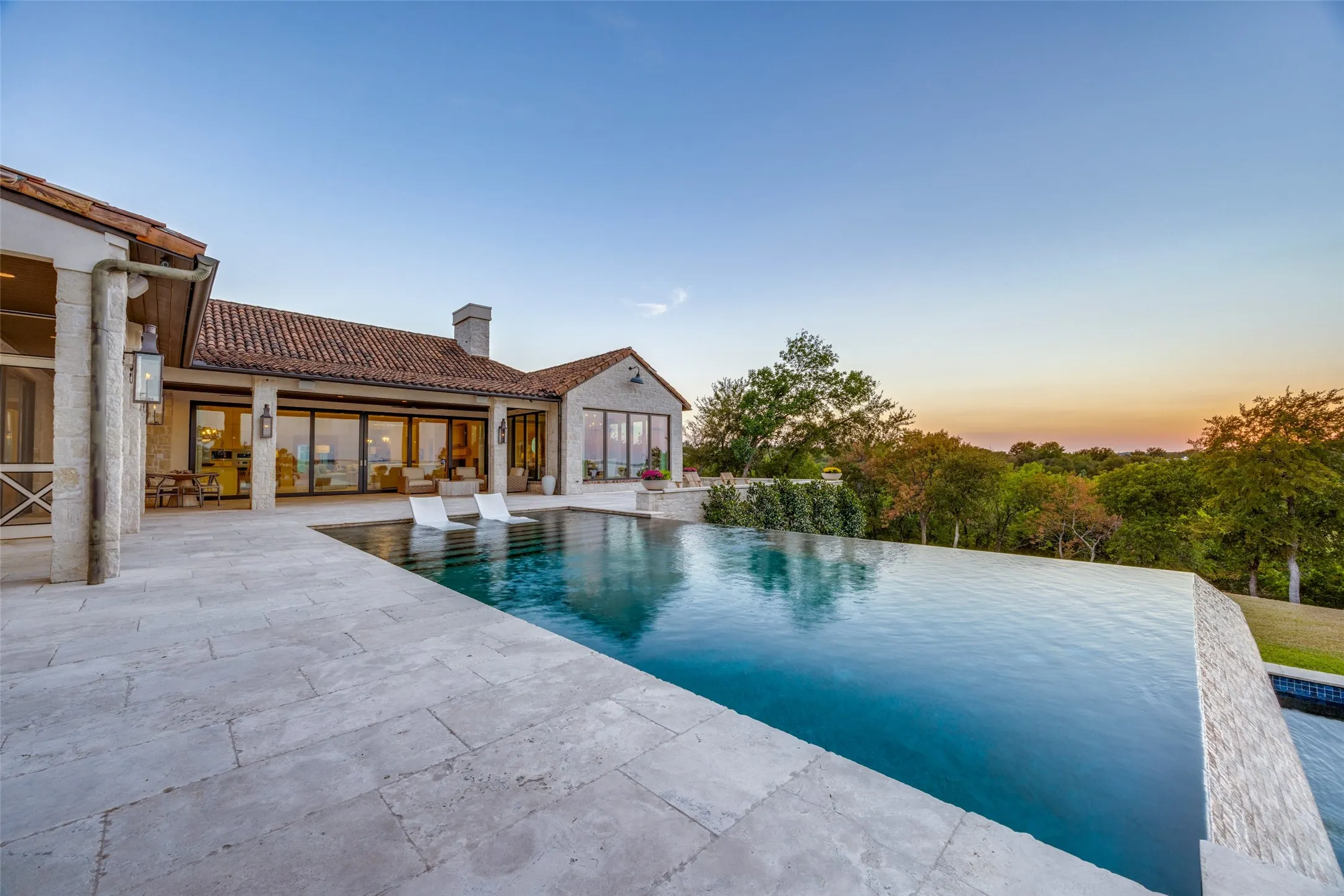Infinity-edge swimming pool with a stone patio, terracotta tile roof, and multiple large glass panel doors
