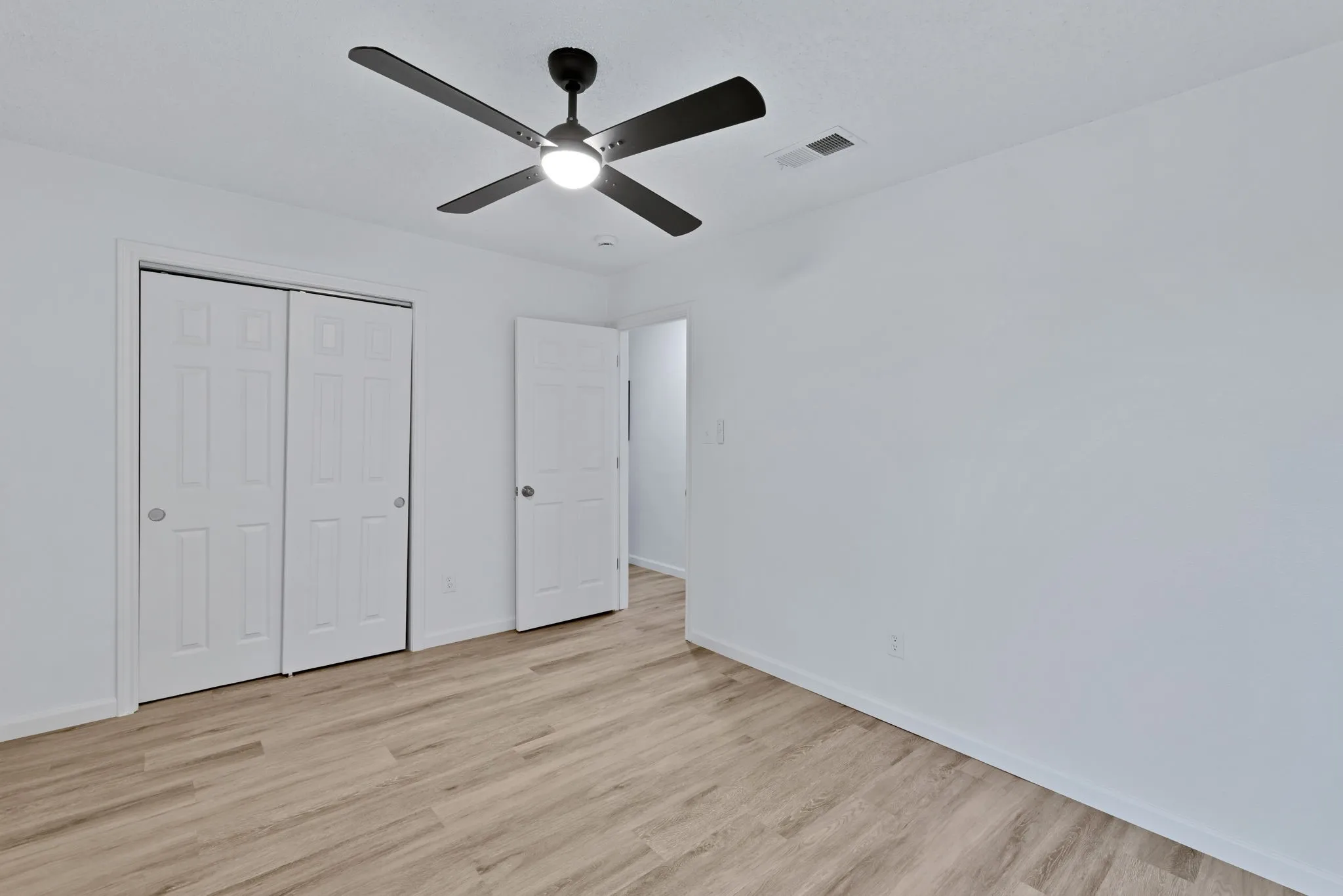 Light-toned wood-finish flooring, white walls, white paneled bifold closet doors, and a black ceiling fan with integrated lighting