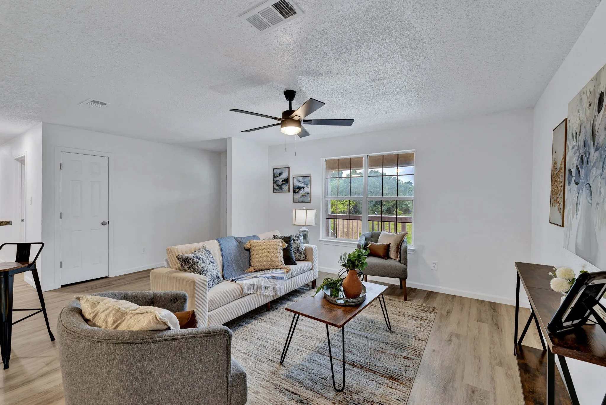 Bright, open living area featuring hardwood flooring, a ceiling fan with integrated lighting, and large windows framed with natural light