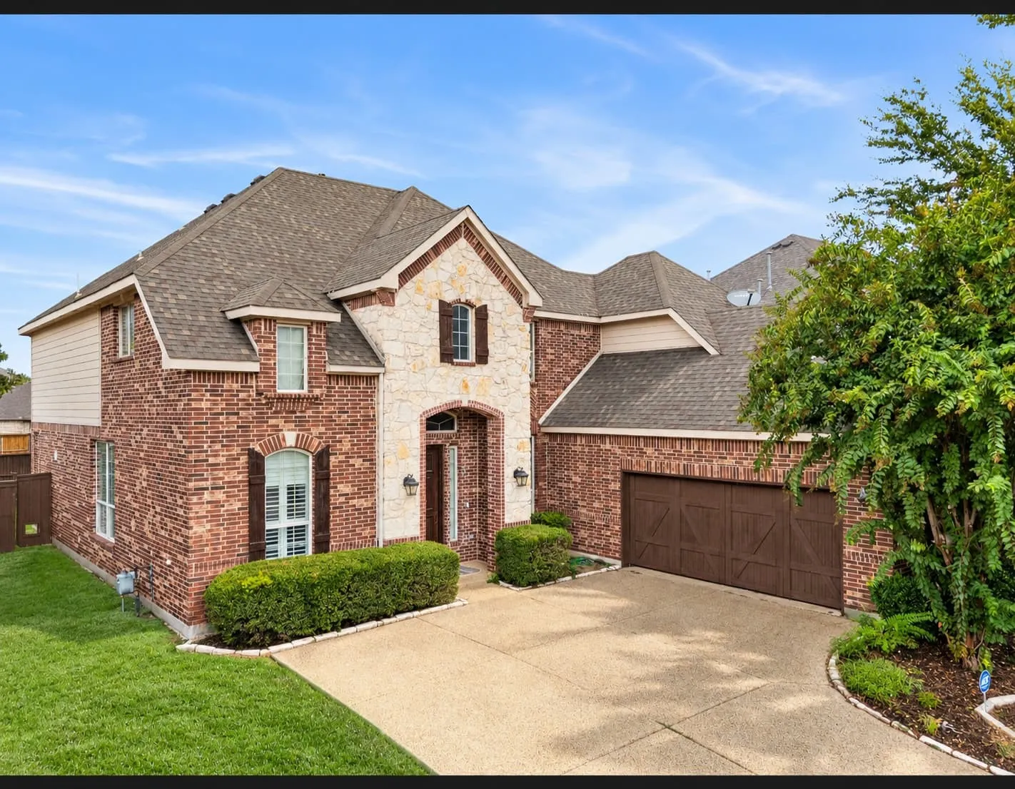 Brick and stone exterior with a peaked roofline, attached garage, concrete driveway, and manicured landscaping