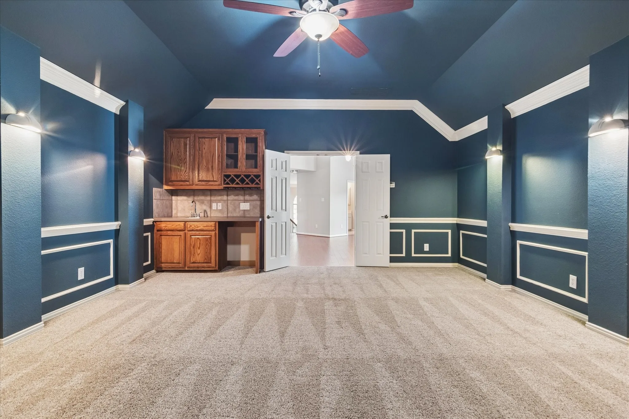 Built-in wet bar with wood cabinetry and tile backsplash, ceiling fan with light fixture, recessed wall paneling, and wall-mounted sconces
