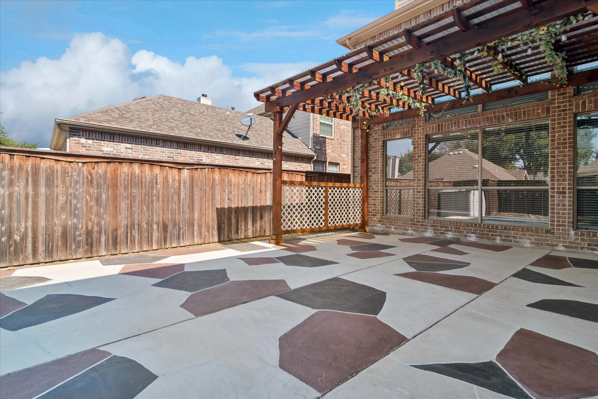 Expansive patio with decorative patterned concrete flooring, a wood pergola structure, and a brick exterior wall with multiple windows