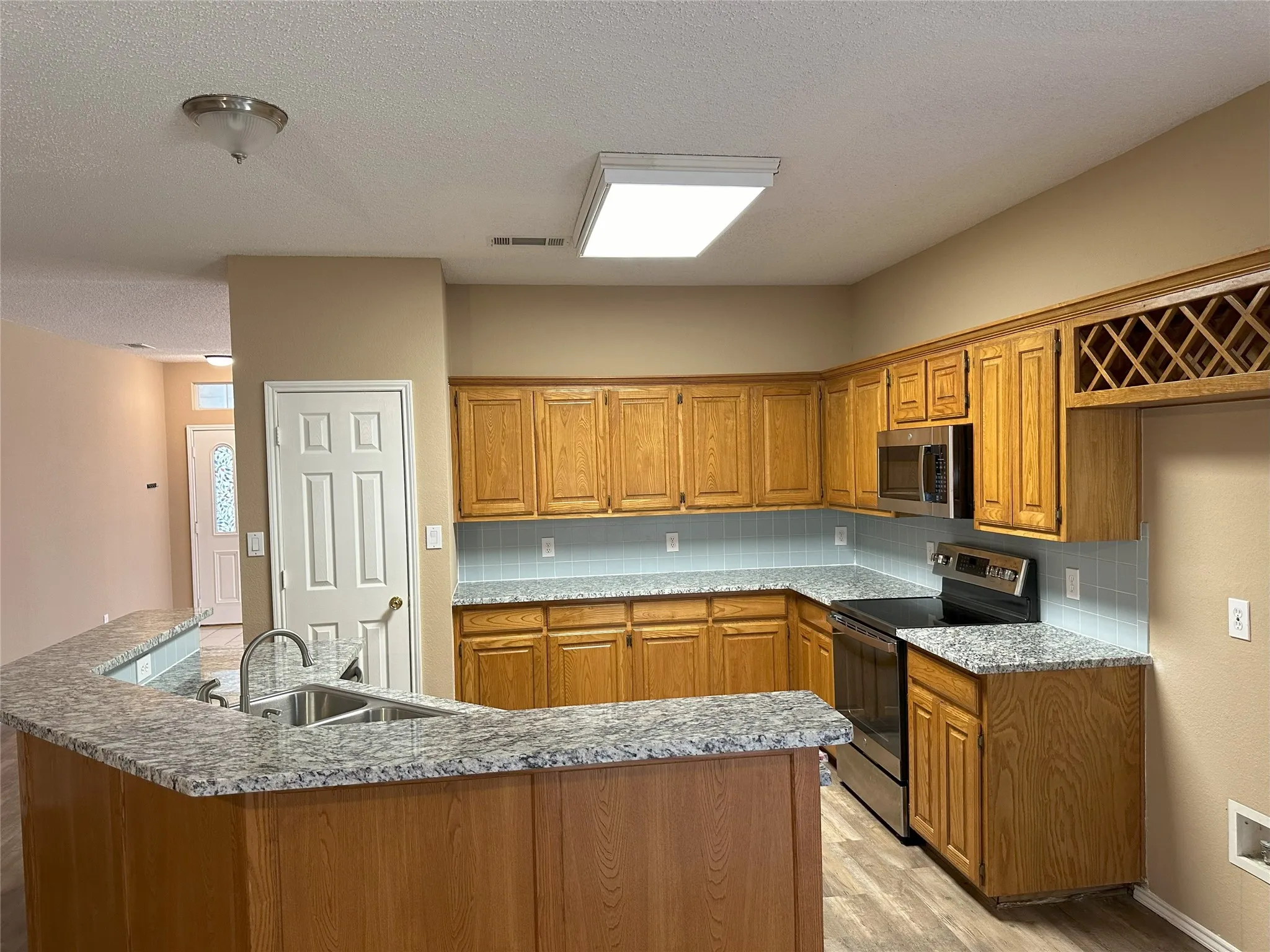 Kitchen featuring a curved granite-finish breakfast bar, stainless steel appliances, light wood cabinetry, a tile backsplash, and wood-finish flooring