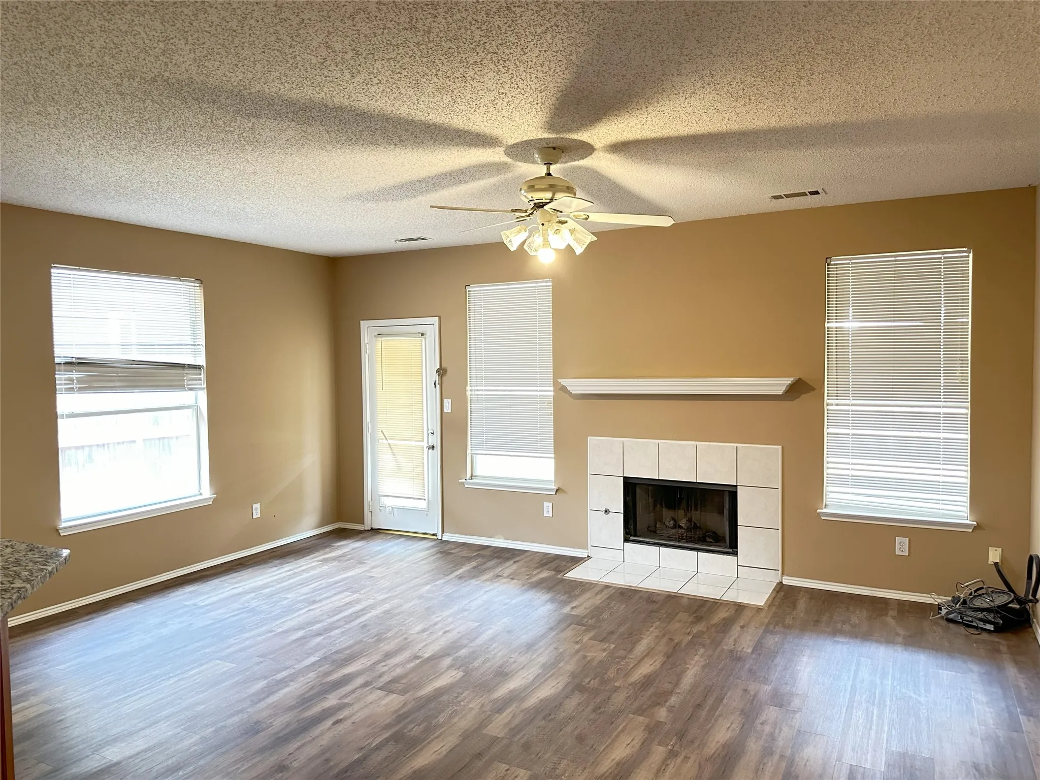 Spacious living area featuring wood-finish flooring, a tiled fireplace with mantel, and multiple windows with blinds
