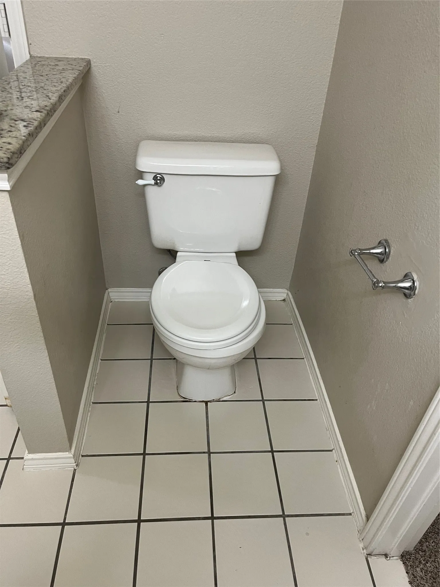 White ceramic toilet with chrome lever, light-colored tile flooring, and a granite-finish countertop