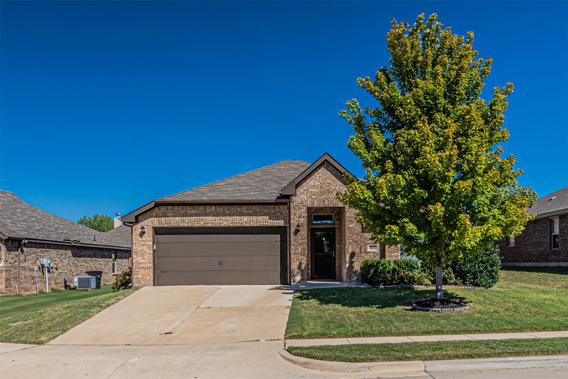 Brick exterior with an attached garage, landscaped front yard, concrete driveway, and a prominent deciduous tree
