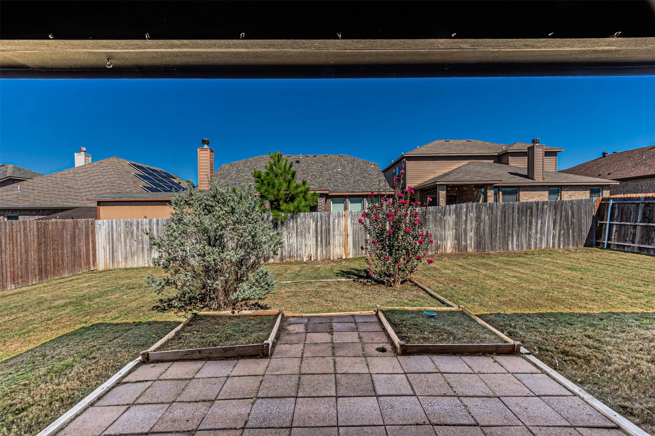 Paved patio area with two raised garden beds, surrounded by a grassy lawn and a wood fence