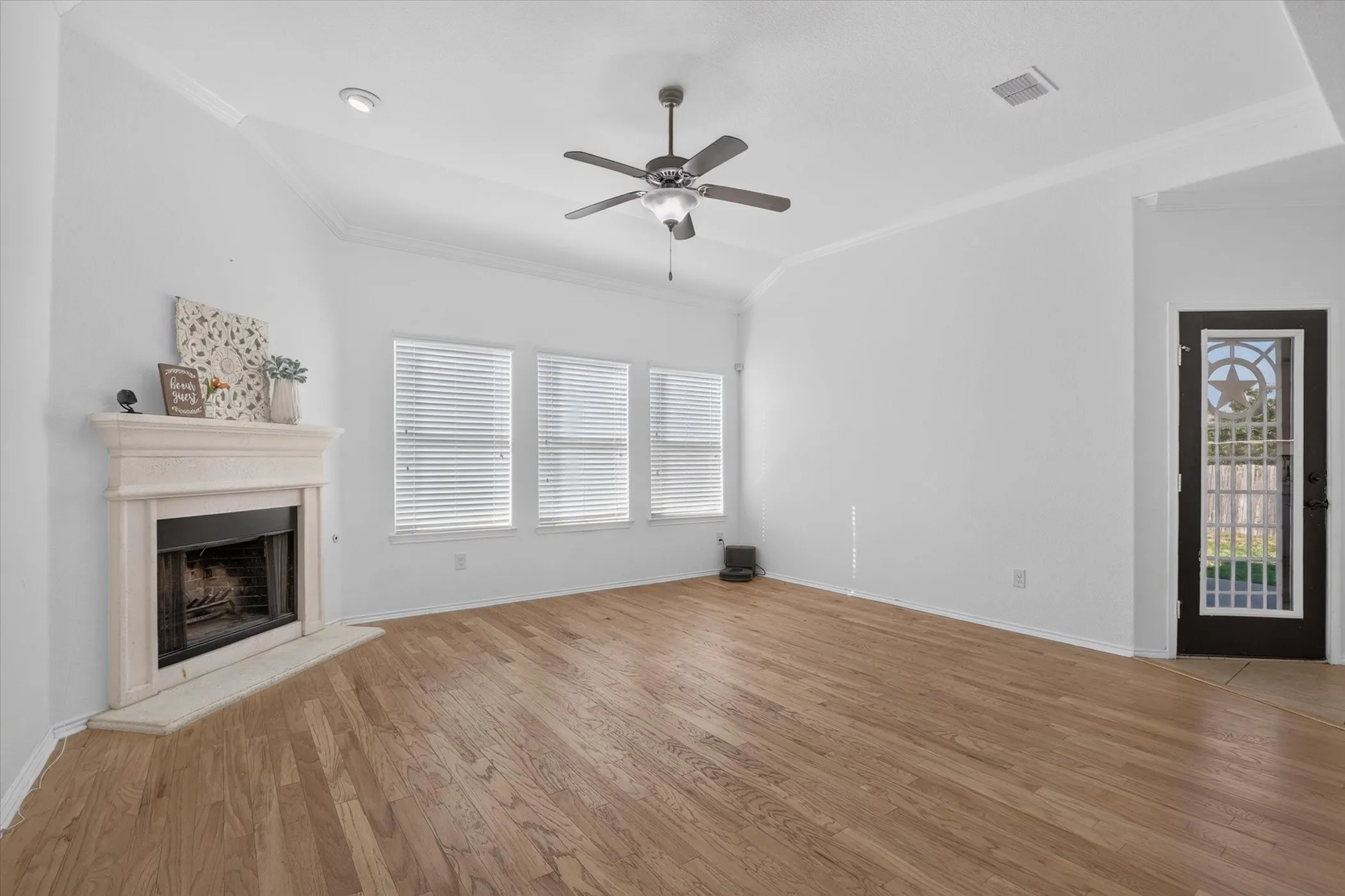 Vaulted ceiling with a ceiling fan, hardwood flooring, stone fireplace with a hearth, and multiple windows with blinds