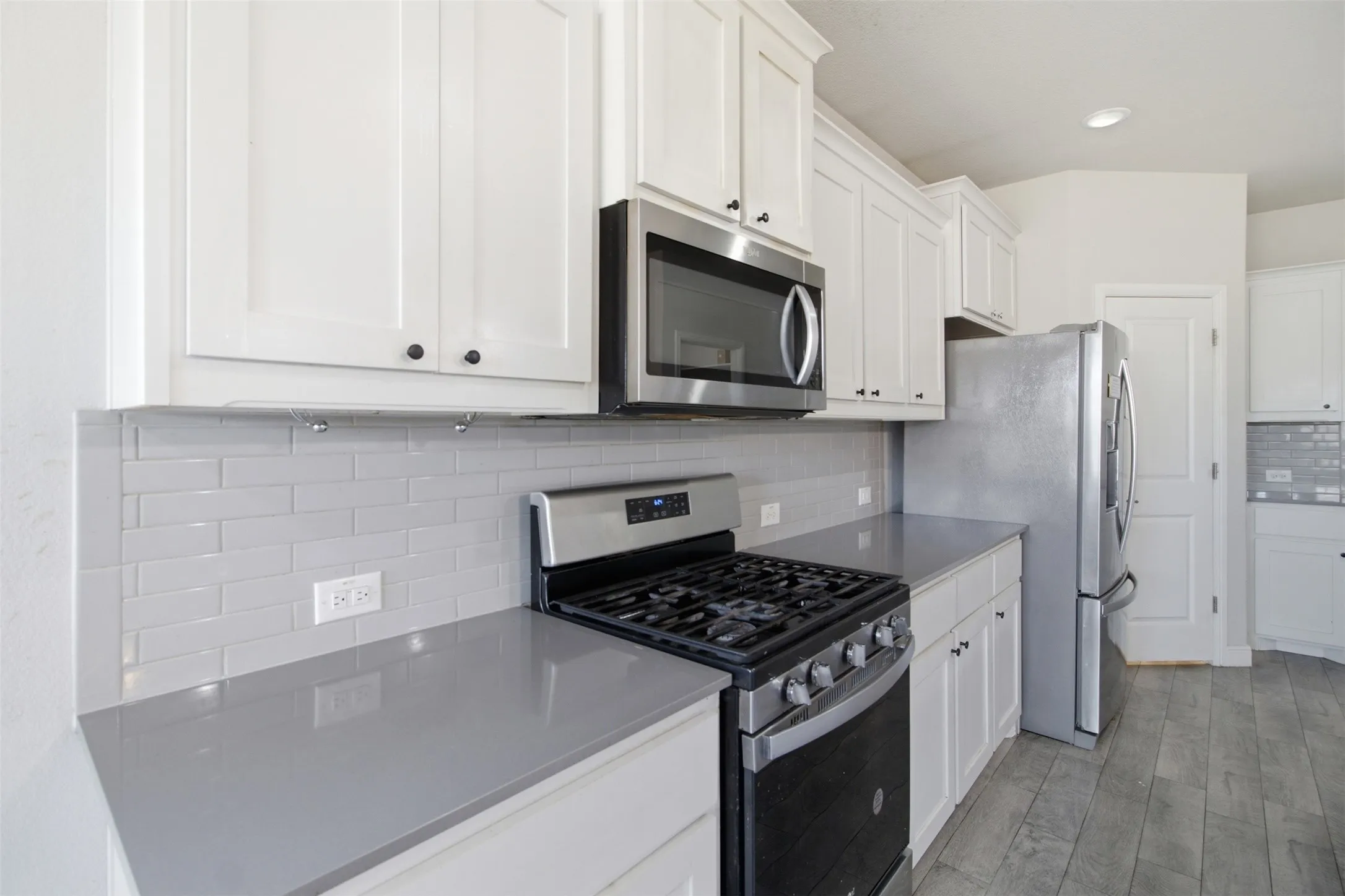 White shaker cabinetry, grey subway tile backsplash, stainless steel appliances, grey countertops, and wood-finish flooring