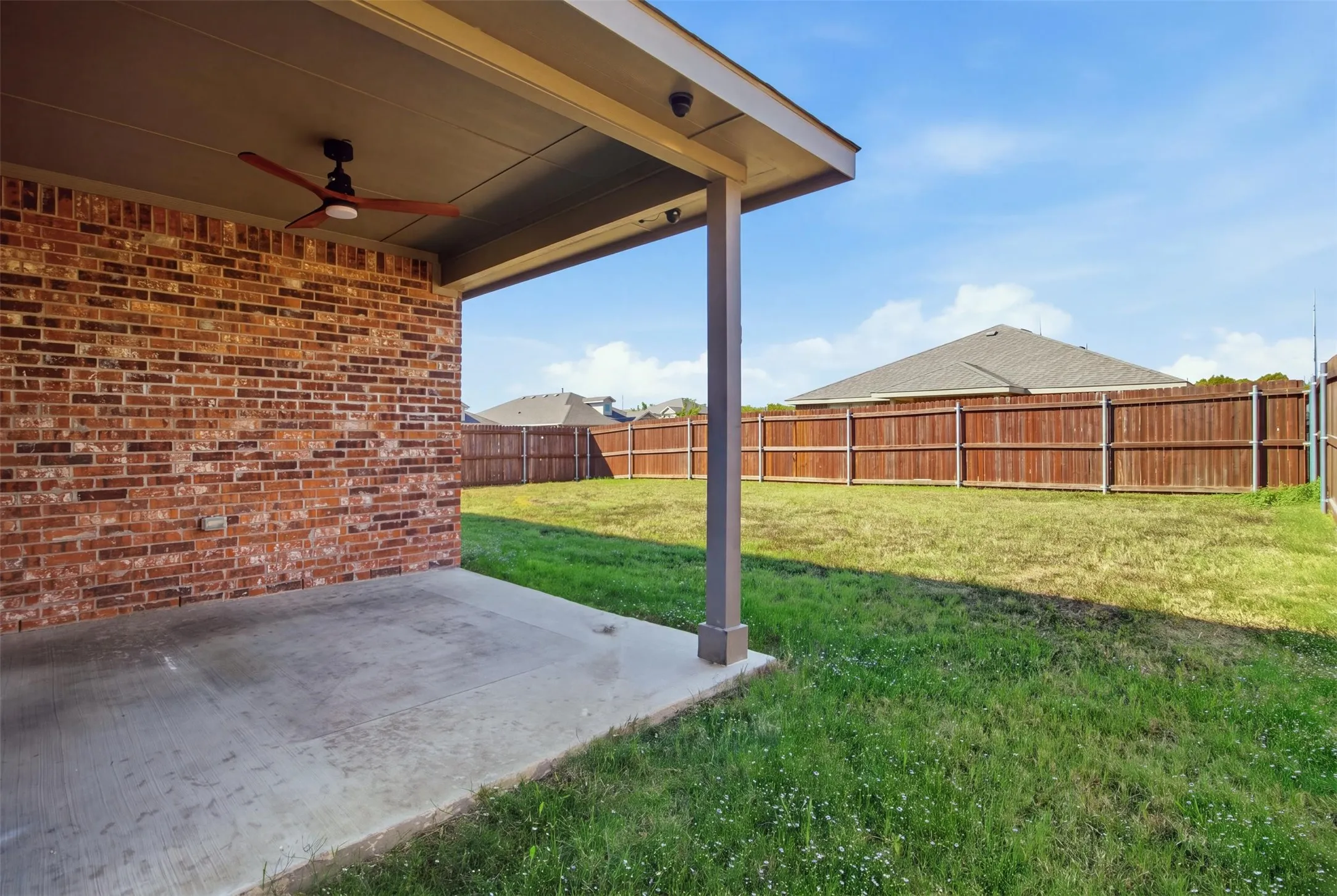 Covered patio with a ceiling fan, brick exterior, concrete flooring, and an expansive grassy yard with a wood privacy fence