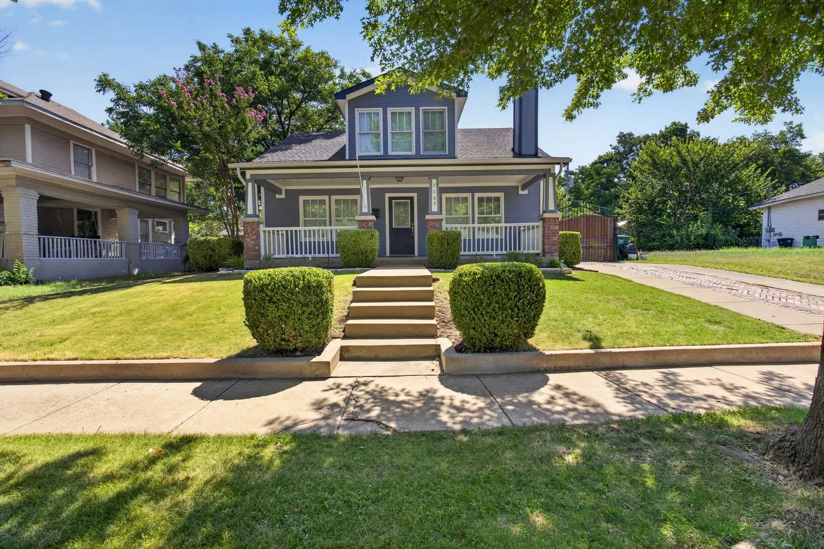 Two-story residence featuring a covered front porch with white railings, a front yard with mature landscaping, and a paved driveway