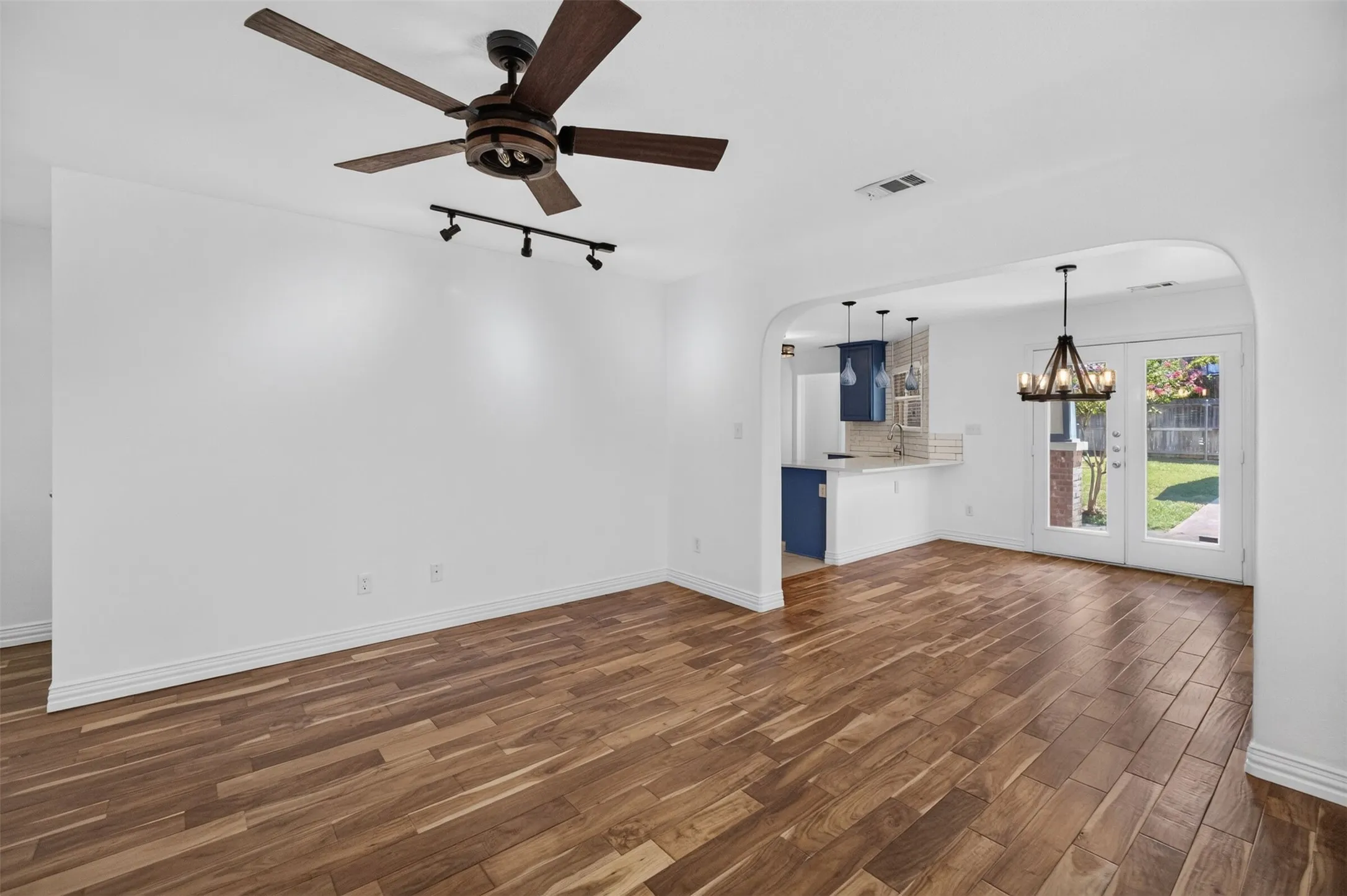 Wood-finish flooring throughout, ceiling fan with wood-tone blades, track lighting, arched doorway, and French doors leading to a backyard