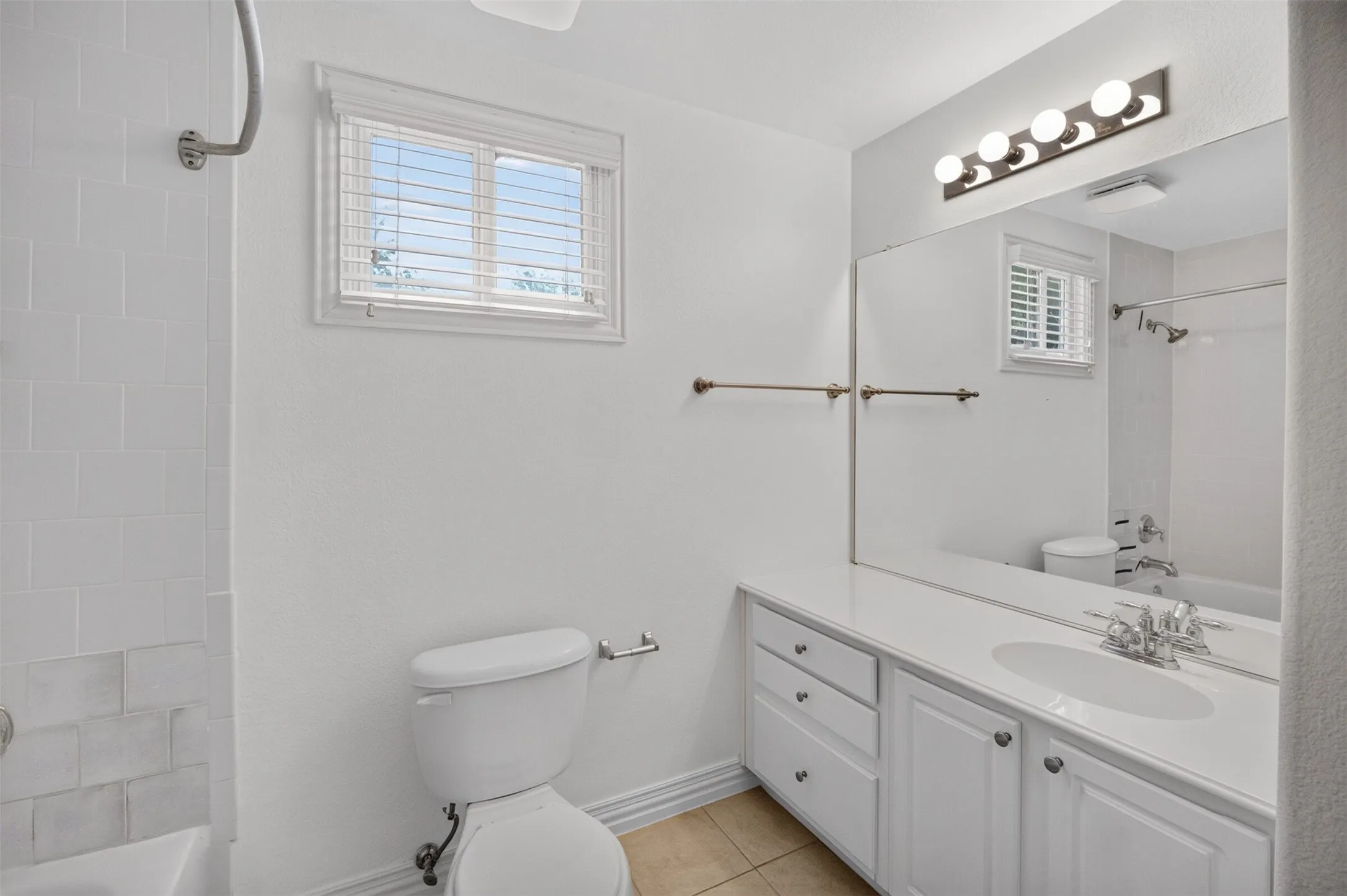 White vanity with integrated sink and chrome faucet, large vanity mirror, four-bulb vanity light fixture, window with blinds, and white subway tile bath surround