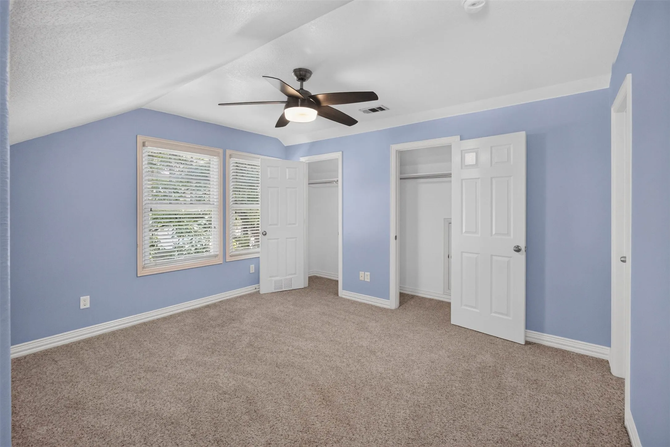 Carpeted flooring, vaulted ceiling, dual windows with blinds, ceiling fan with integrated lighting, and multiple closet storage