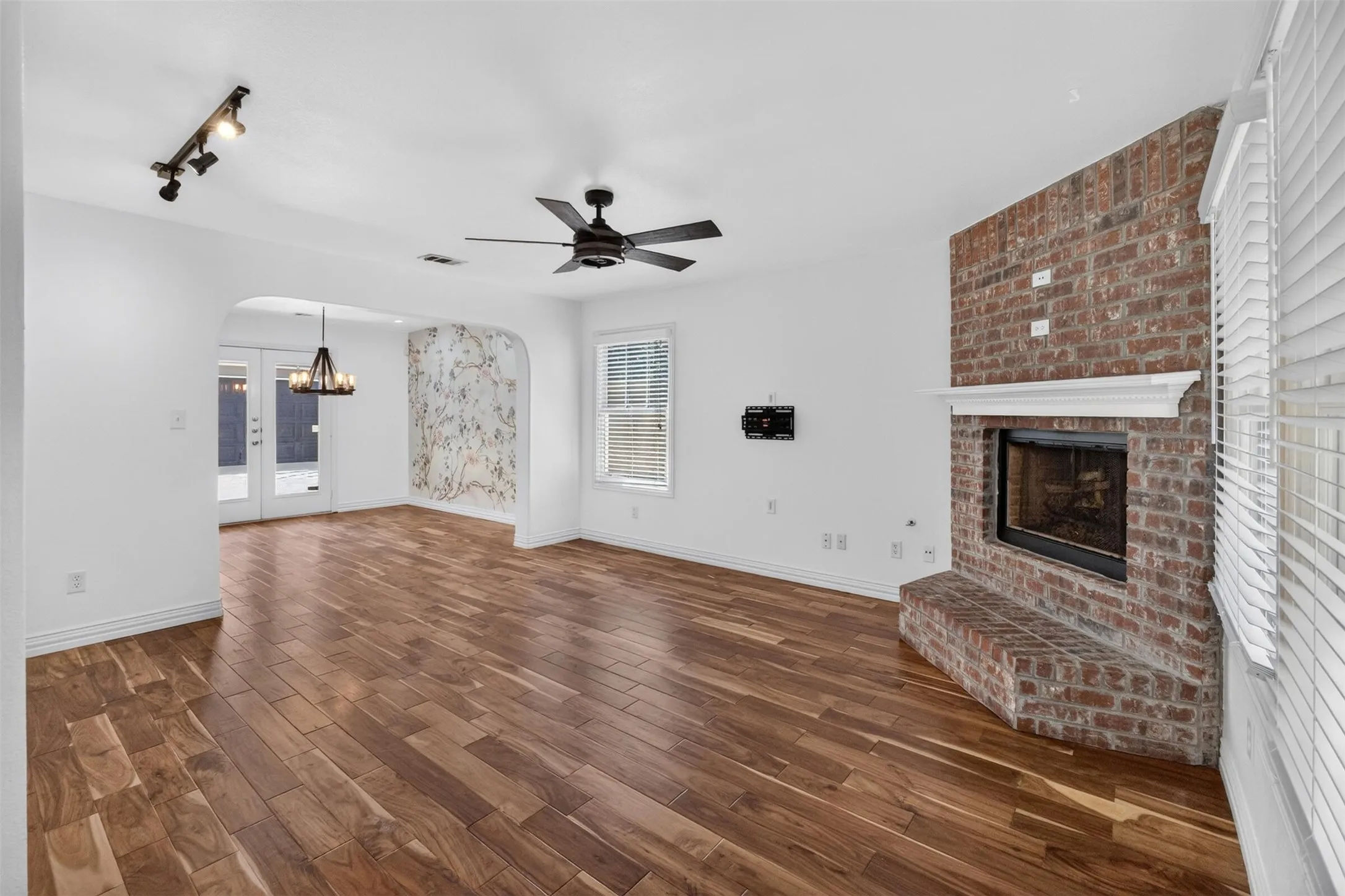 Wood-finish flooring, brick fireplace with a white mantel, ceiling fan, track lighting, and arched doorway
