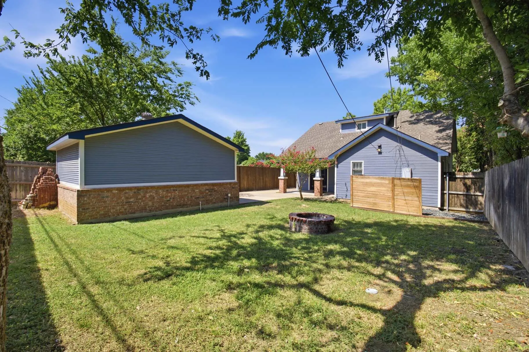 Expansive grassy yard with a brick fire pit, wood fence, and a separate structure featuring horizontal siding over a brick base
