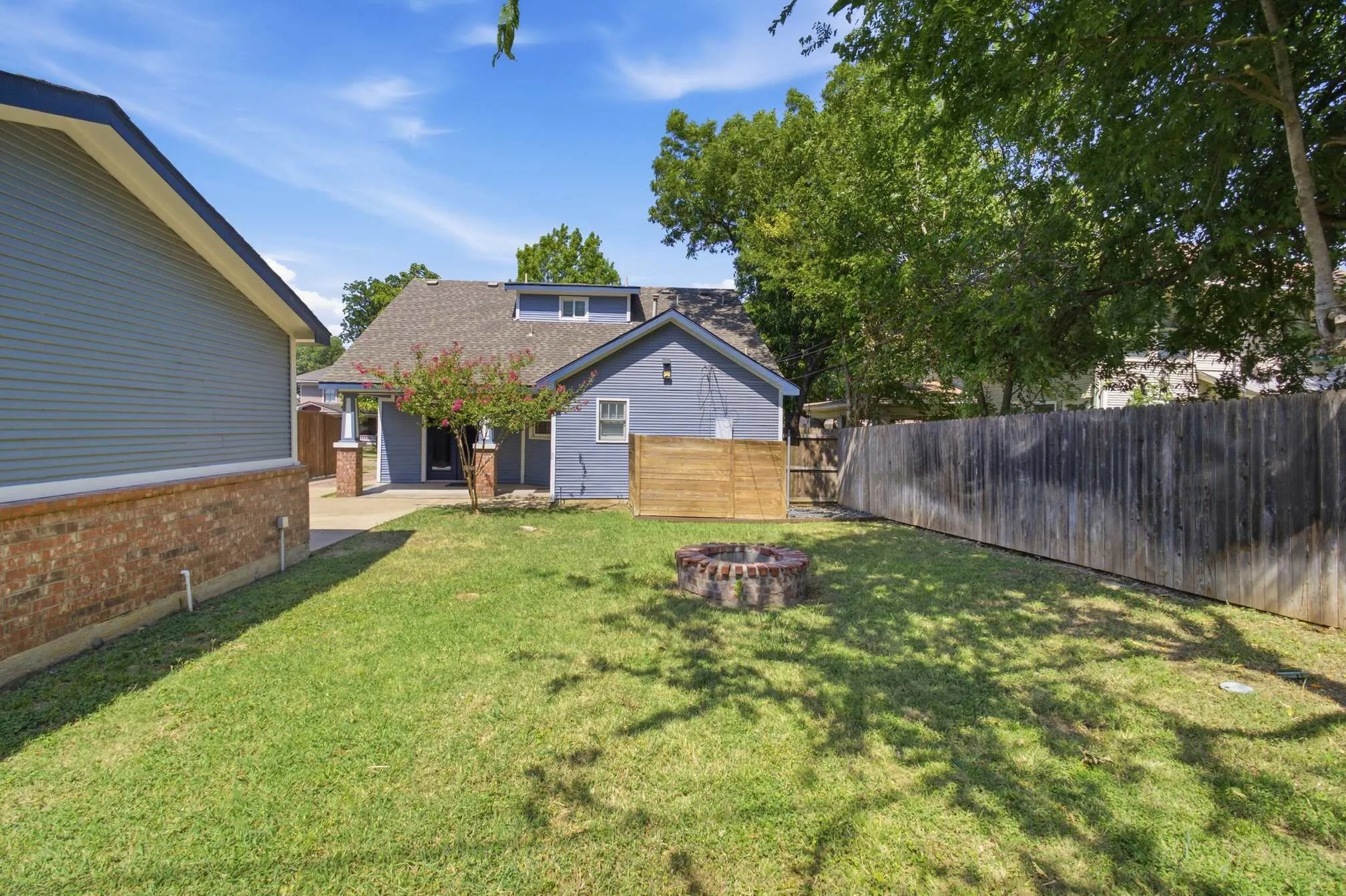 Fenced backyard with a lawn, a brick-ringed fire pit, and mature trees