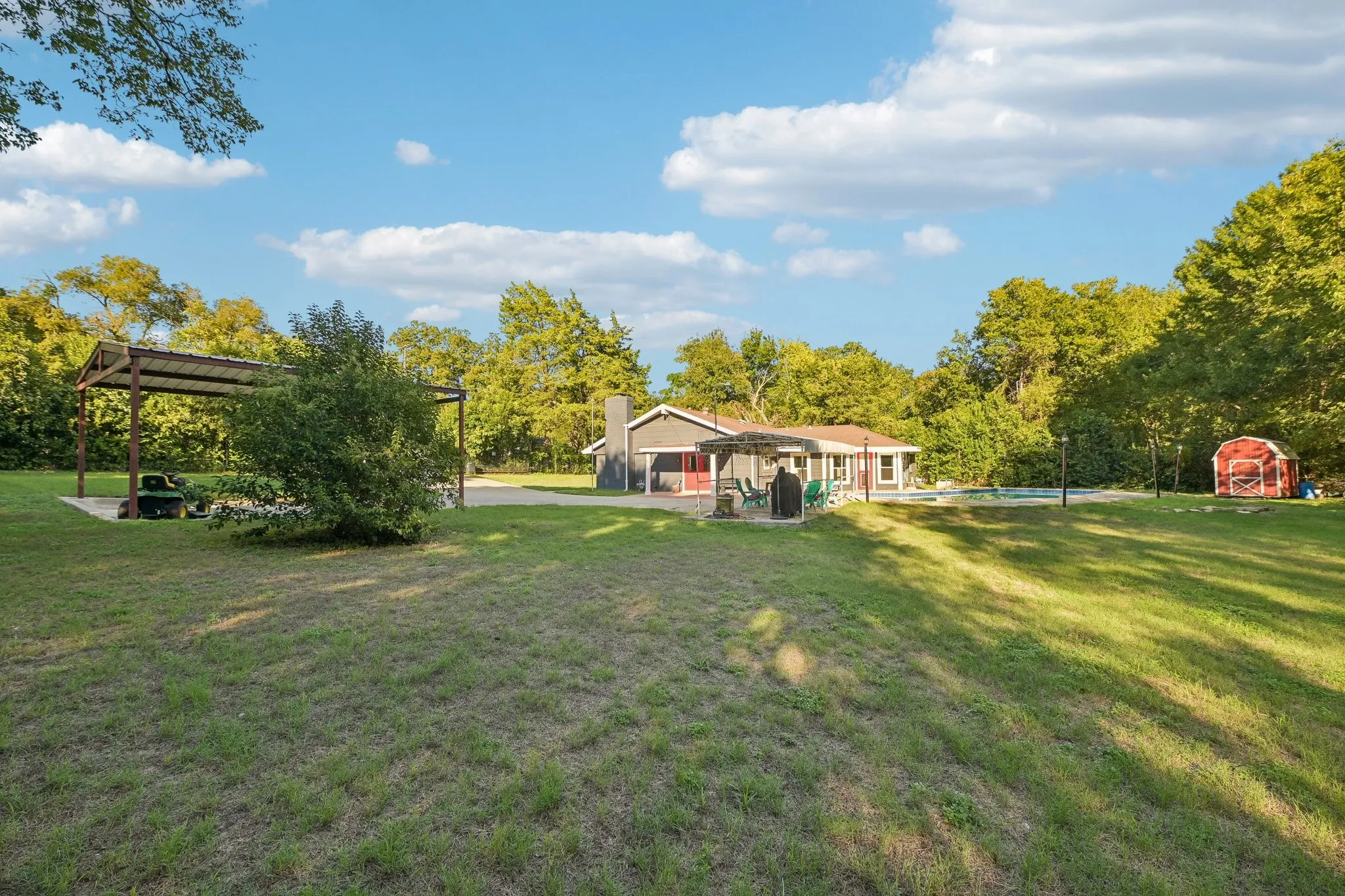 Expansive lawn with a covered outdoor structure, mature trees, a red storage shed, and a visible pool