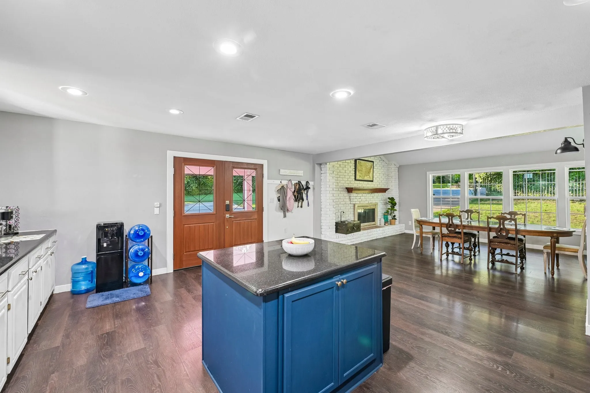 Wood-finish flooring throughout, featuring a kitchen island with storage and a dark countertop, white perimeter cabinetry, and an exposed brick fireplace