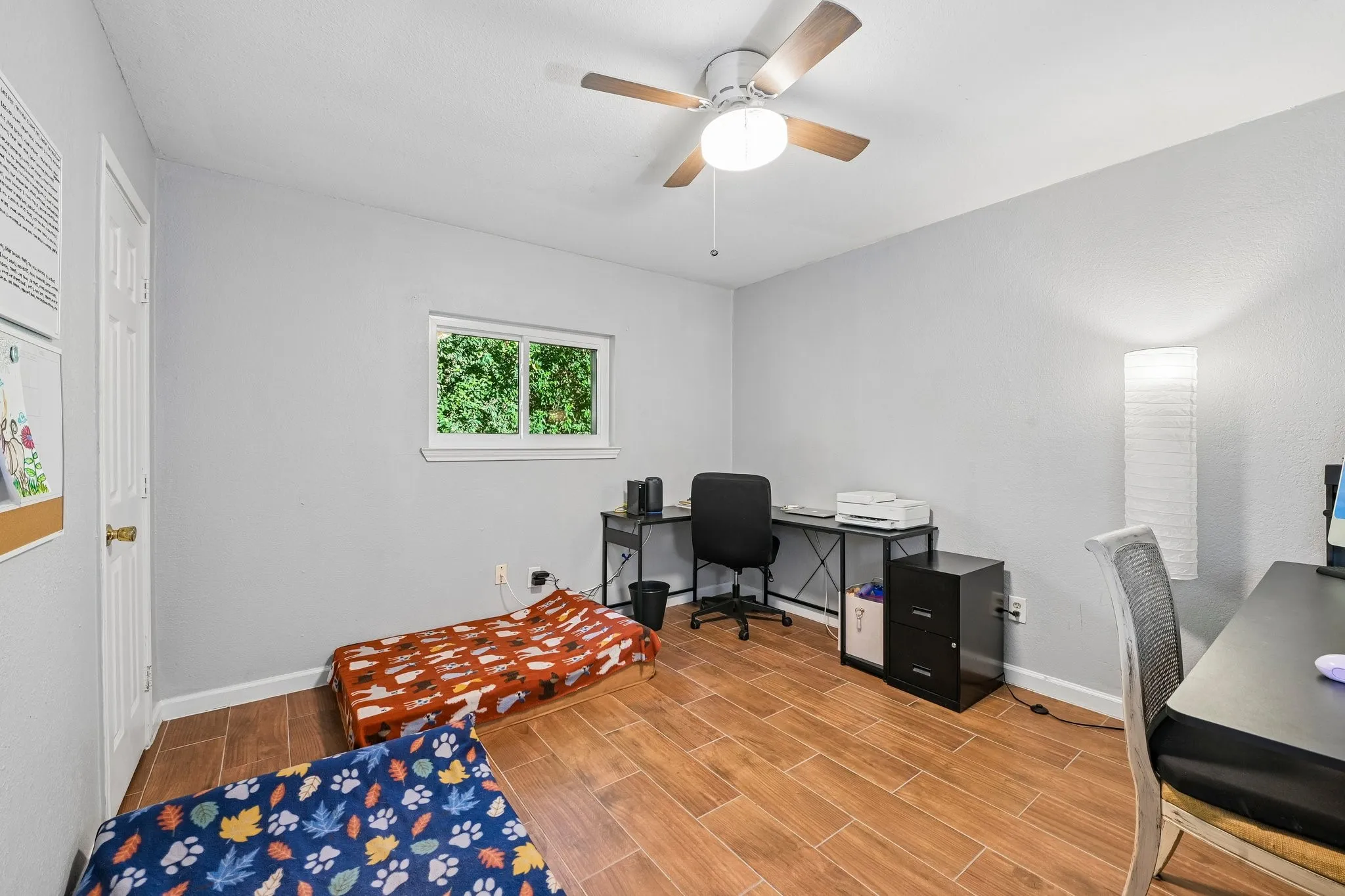 Wood-finish tile flooring throughout, featuring a single window, a ceiling fan with integrated lighting, and light gray wall paint
