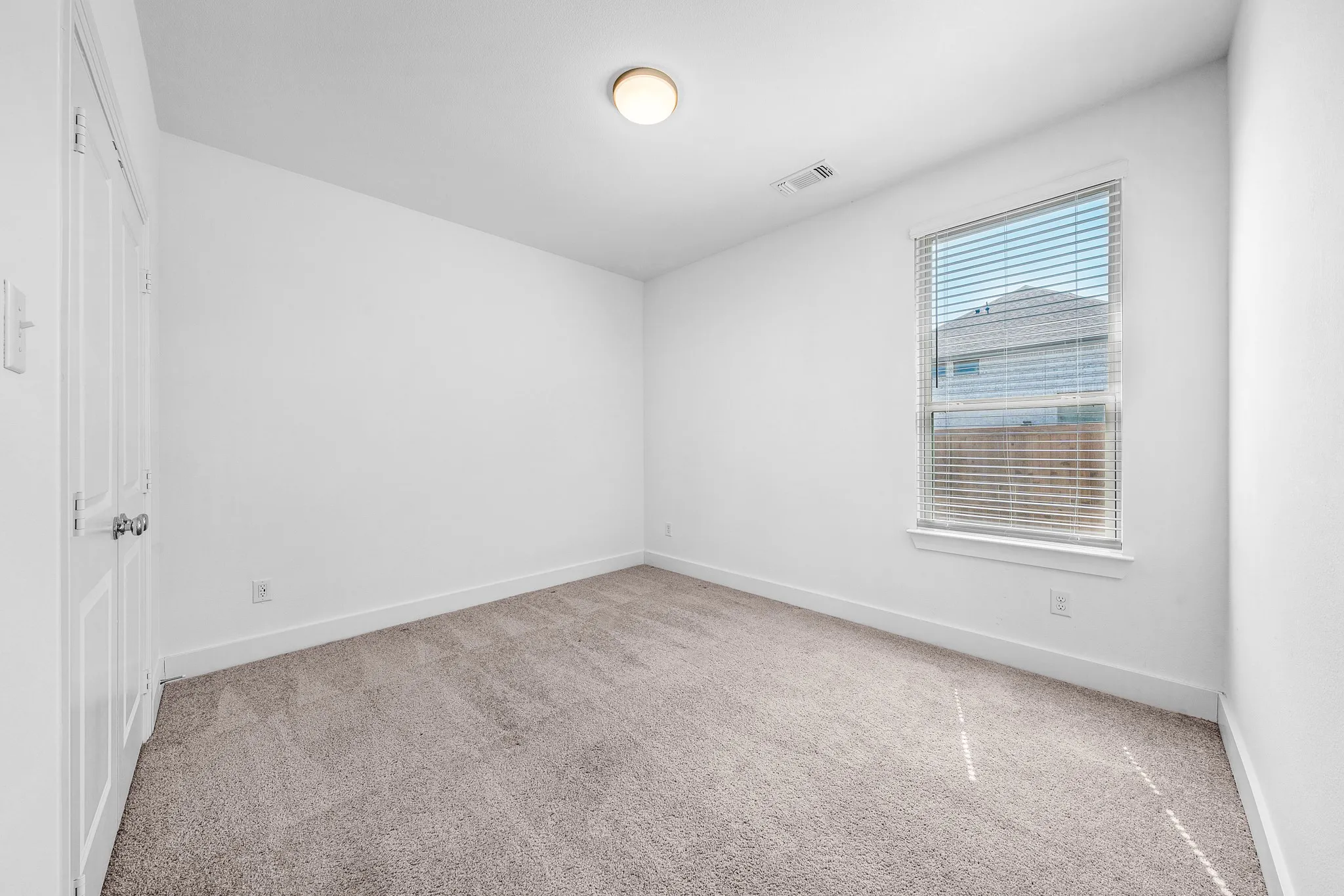 Neutral tone carpet, white painted walls, single window with blinds, ceiling-mounted light fixture, paneled door with knob