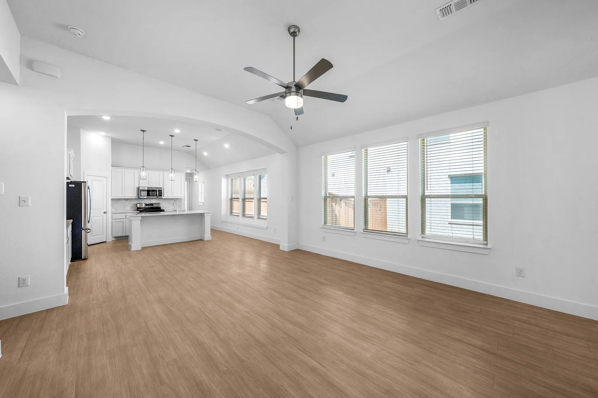 Vaulted ceiling with a ceiling fan, wood-finish flooring, multiple windows, and an arched entryway to the kitchen
