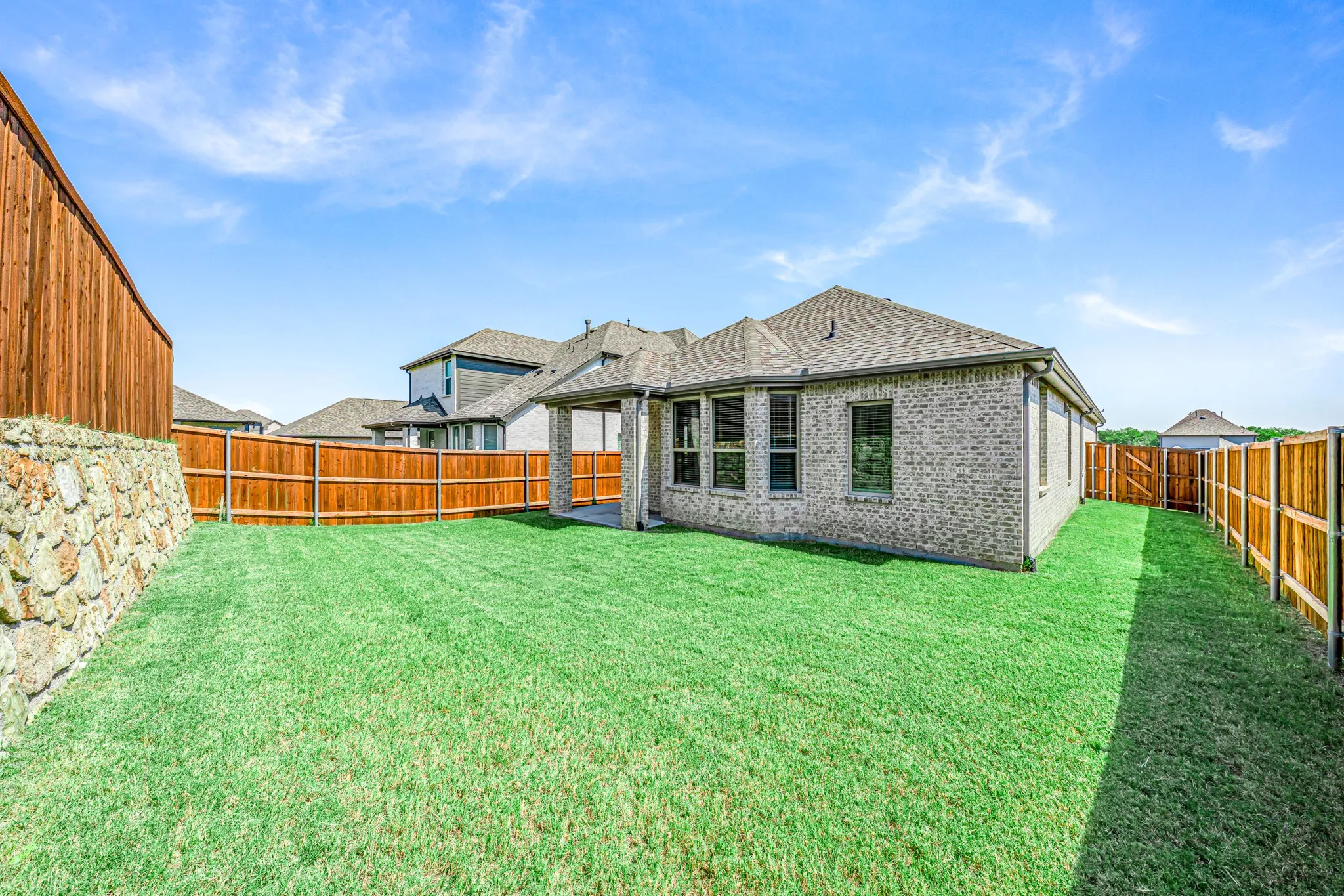 Expansive fenced backyard with a stone retaining wall, covered patio, and brick exterior
