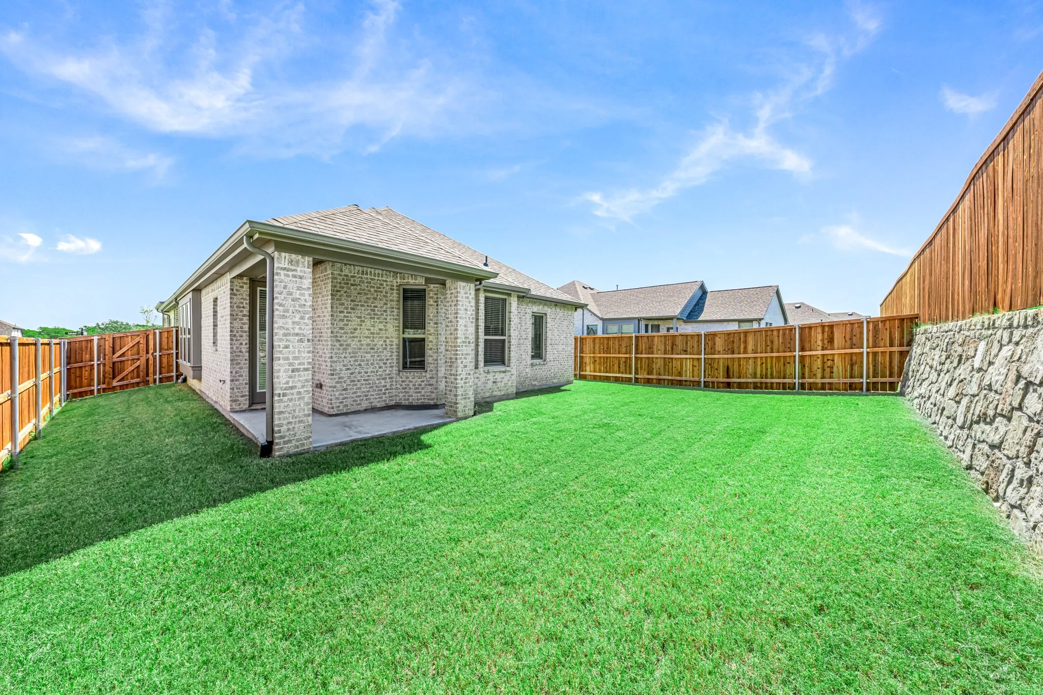 Expansive backyard featuring a lush lawn, privacy fencing, a stone retaining wall, and a covered patio with a brick exterior