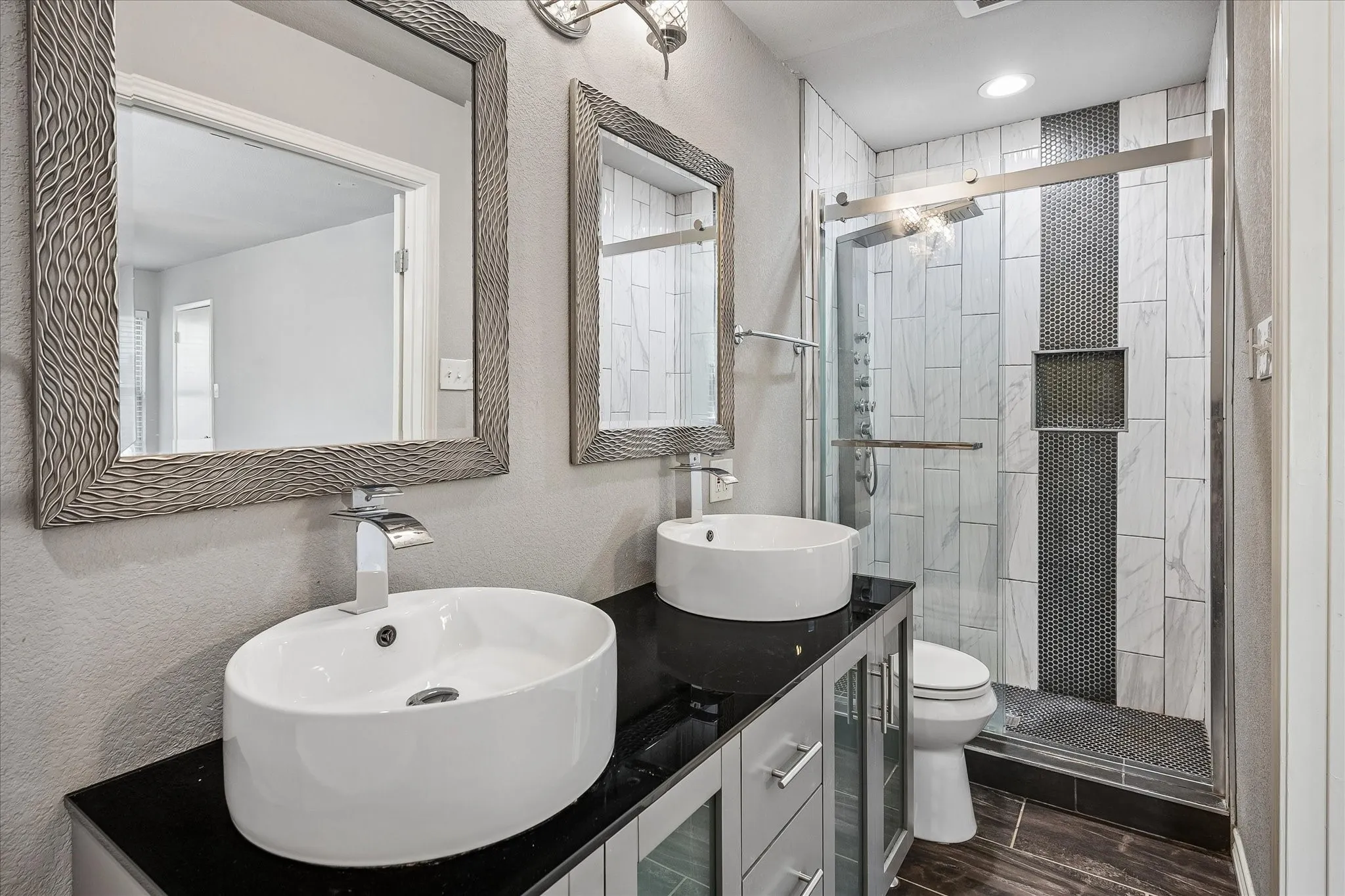Dual vessel sinks set on a black countertop, glass-enclosed shower with contrasting tile, wood-finish flooring, and wall-mounted vanity lighting