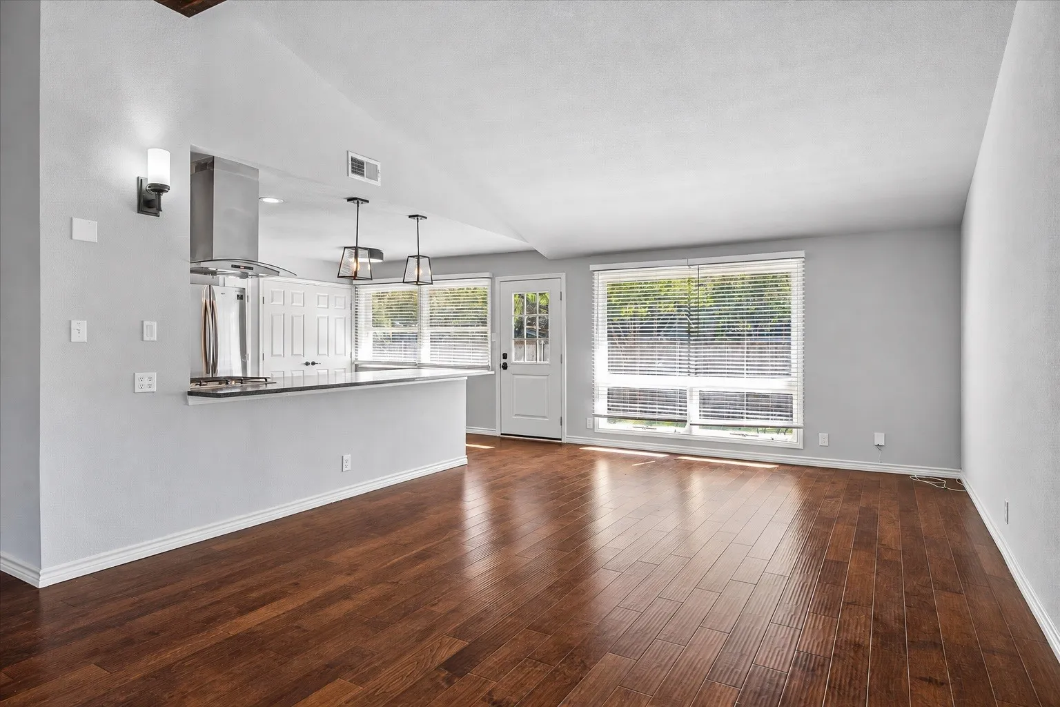 Rich wood-finish flooring, vaulted ceiling, white cabinetry with dark hardware, stainless steel appliances, and large windows with privacy blinds