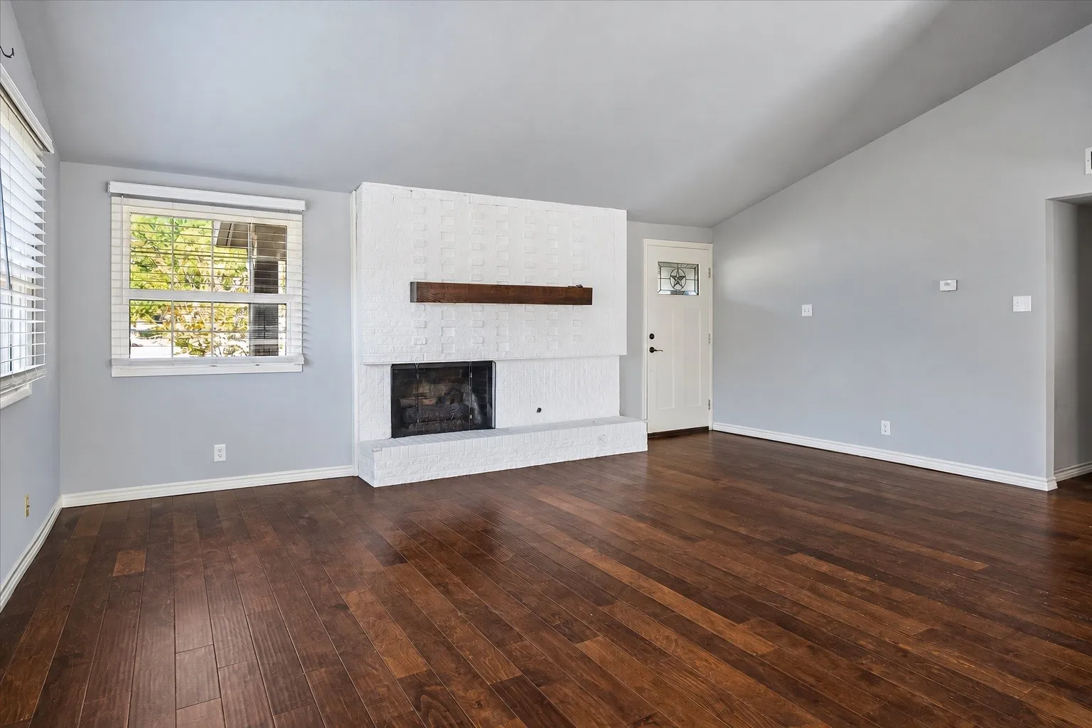 Vaulted ceiling, wood-finish flooring, white brick fireplace with wood mantel, white entry door with decorative glass inlay, and light gray painted walls