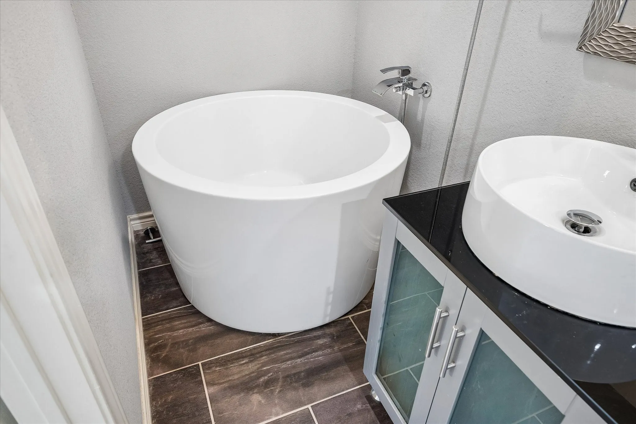 Freestanding soaking tub, vessel sink, vanity with frosted glass doors, dark countertop, and tile flooring