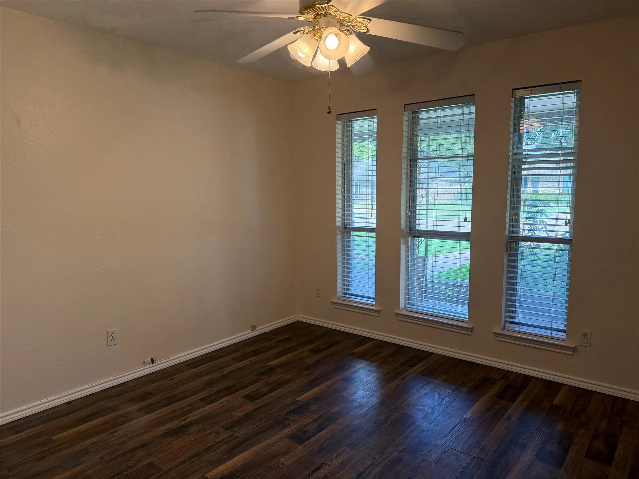 Wood-finish flooring, three vertical windows with blinds, and ceiling fan with integrated lighting