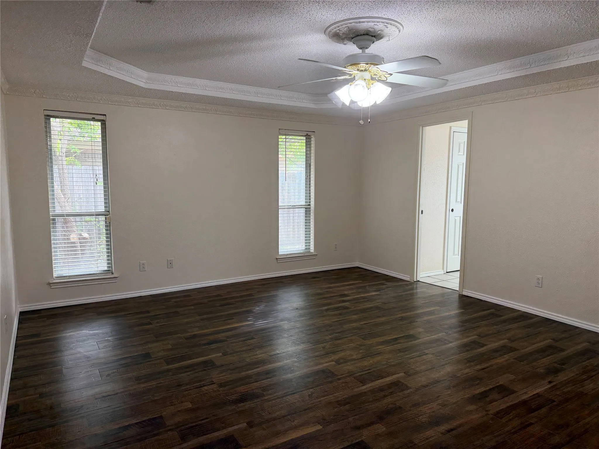 Tray ceiling with crown molding, ceiling fan with light fixture, wood-finish flooring, and two windows with blinds