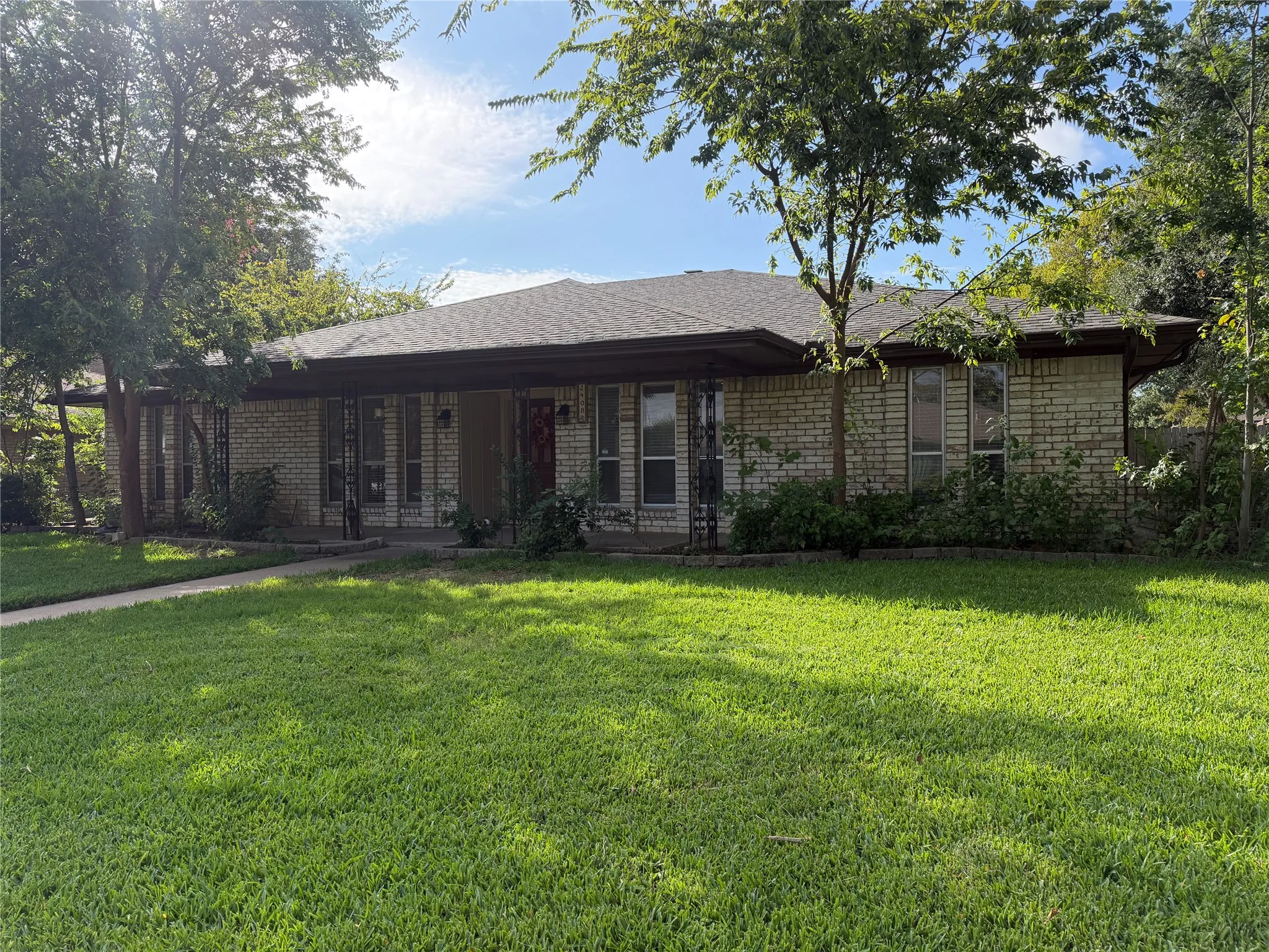 Brick exterior with a covered front porch featuring decorative ironwork, a pitched shingle roof, and a mature tree canopy