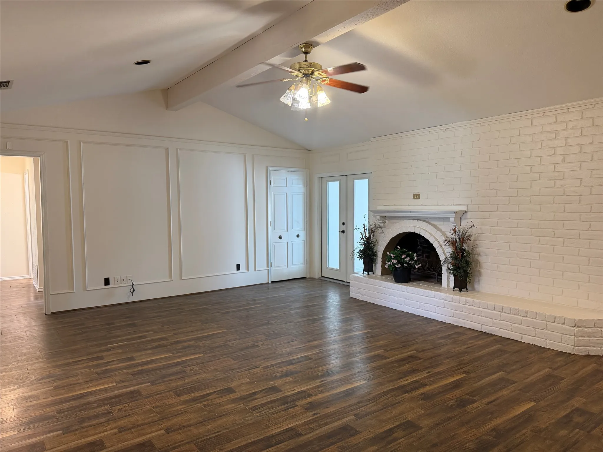 Vaulted ceiling with exposed beam, wood-finish flooring, white brick fireplace with hearth, white paneled wall, and double glass-paneled doors