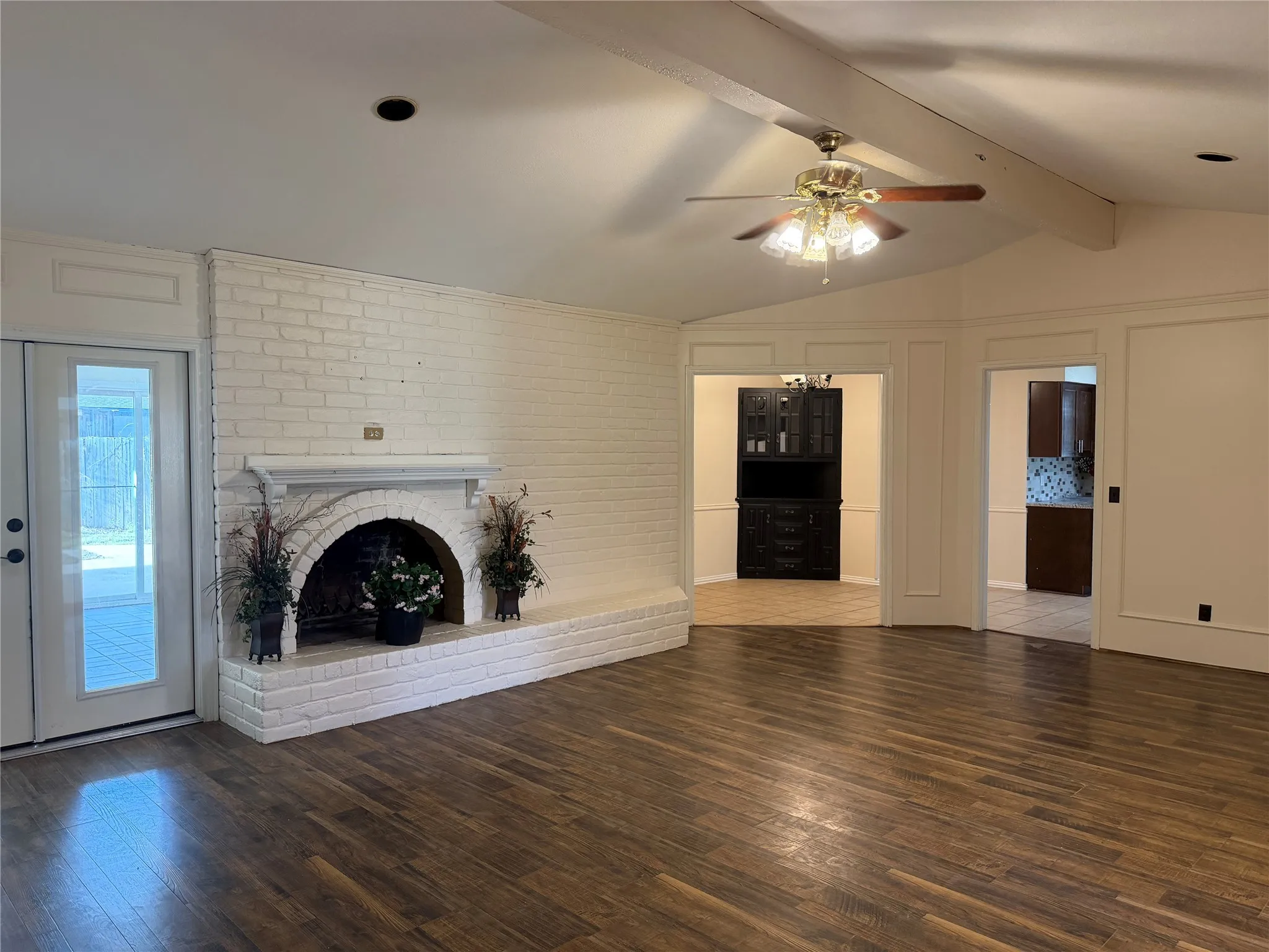 Vaulted ceiling with exposed beam, white brick fireplace, wood-finish flooring, glass-paneled door, and ceiling fan