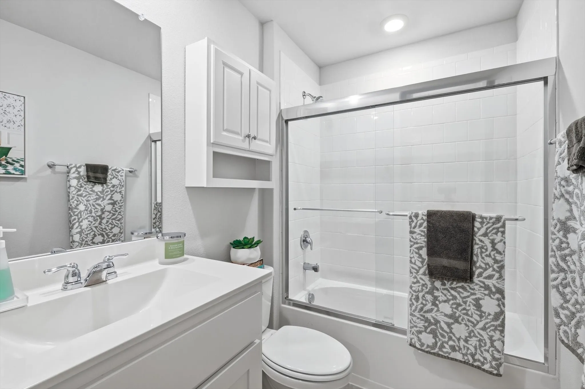 White vanity with integrated sink, chrome faucet, wall-mounted mirror, white shaker-style cabinetry, and a bathtub with a sliding glass enclosure