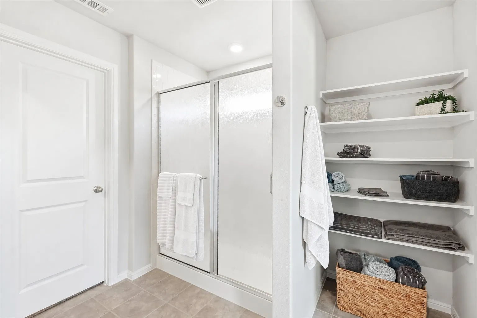 Tiled flooring with a white door, shower enclosure with frosted glass panels, built-in shelving, and recessed lighting