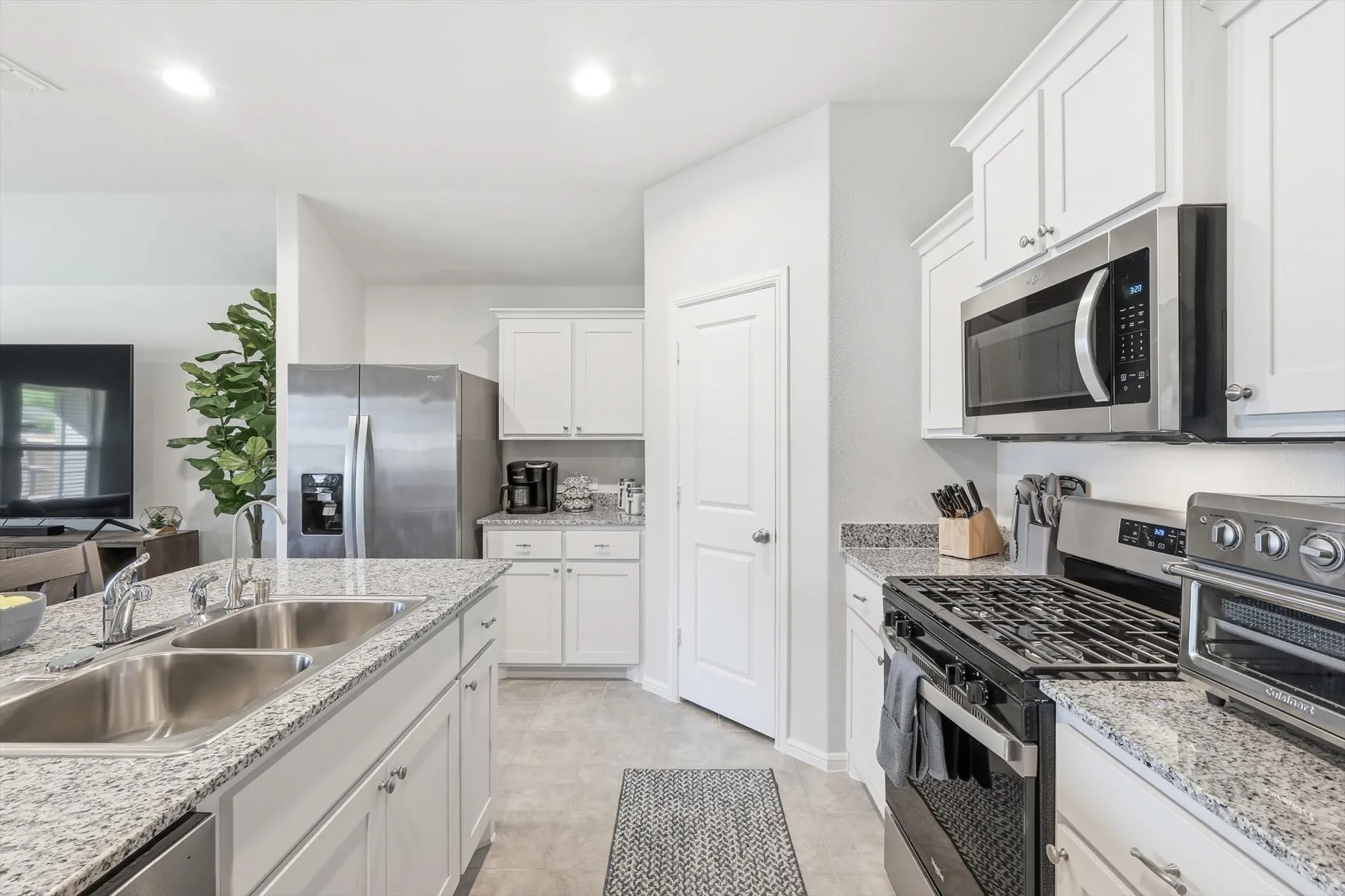 White cabinetry with brushed nickel hardware, stainless steel appliances, dual basin stainless steel sink, light-toned tile flooring, and light-toned speckled countertops