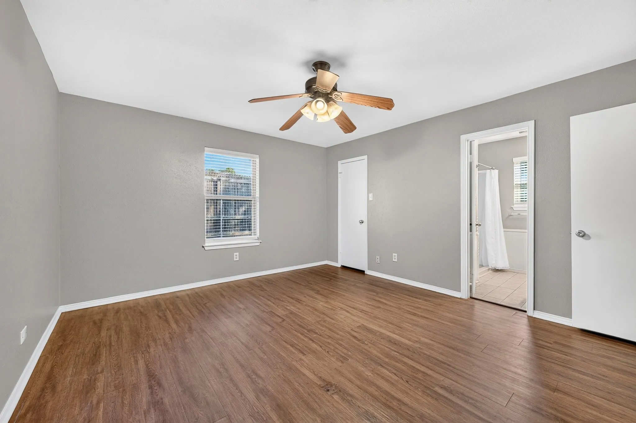 Spacious bedroom featuring warm wood-finish flooring, neutral gray walls, ceiling fan with integrated lighting, and abundant natural light from large window
