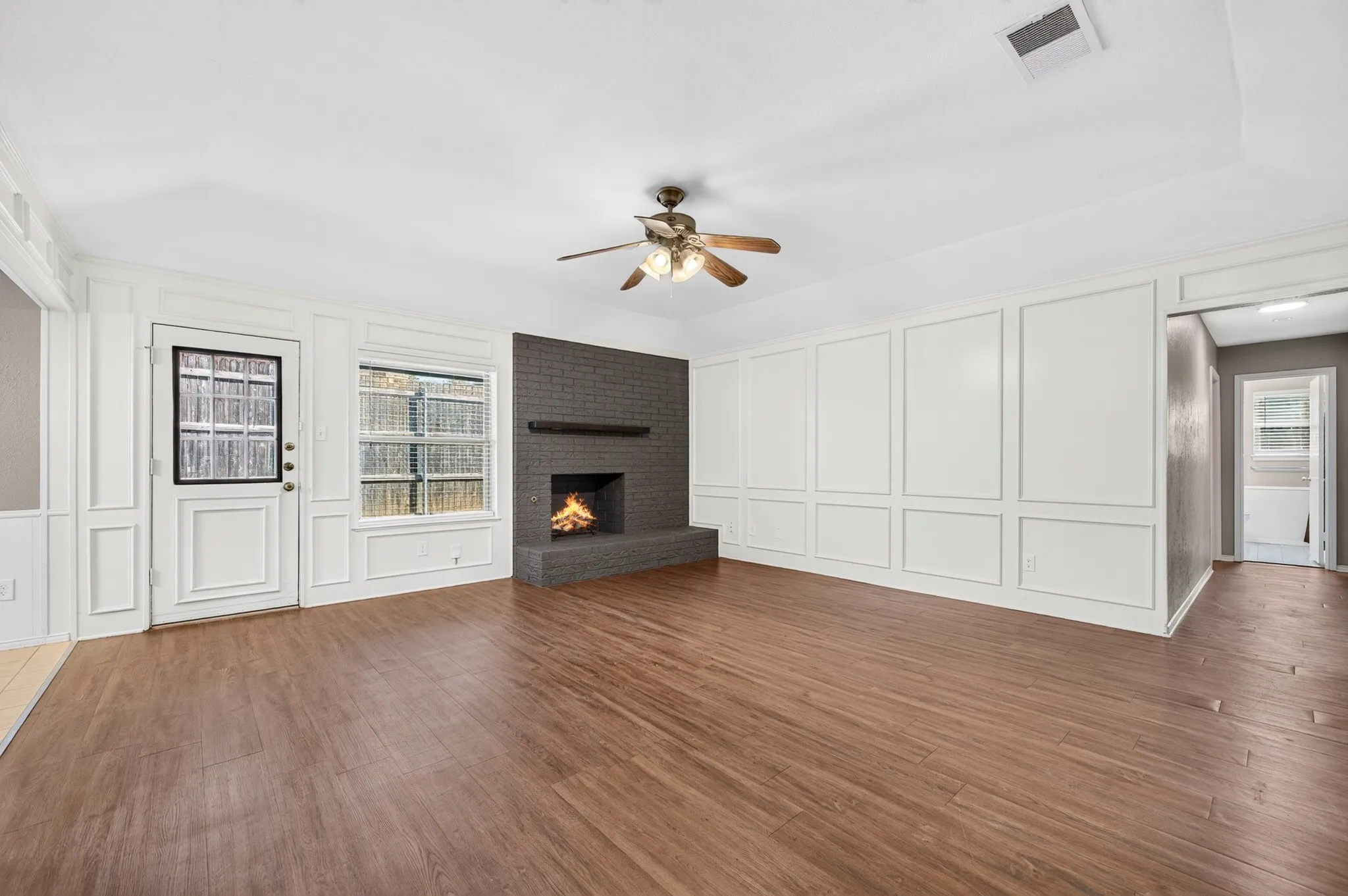 Wood-finish flooring, white paneling, brick fireplace with mantel, ceiling fan, and a glass-paned door