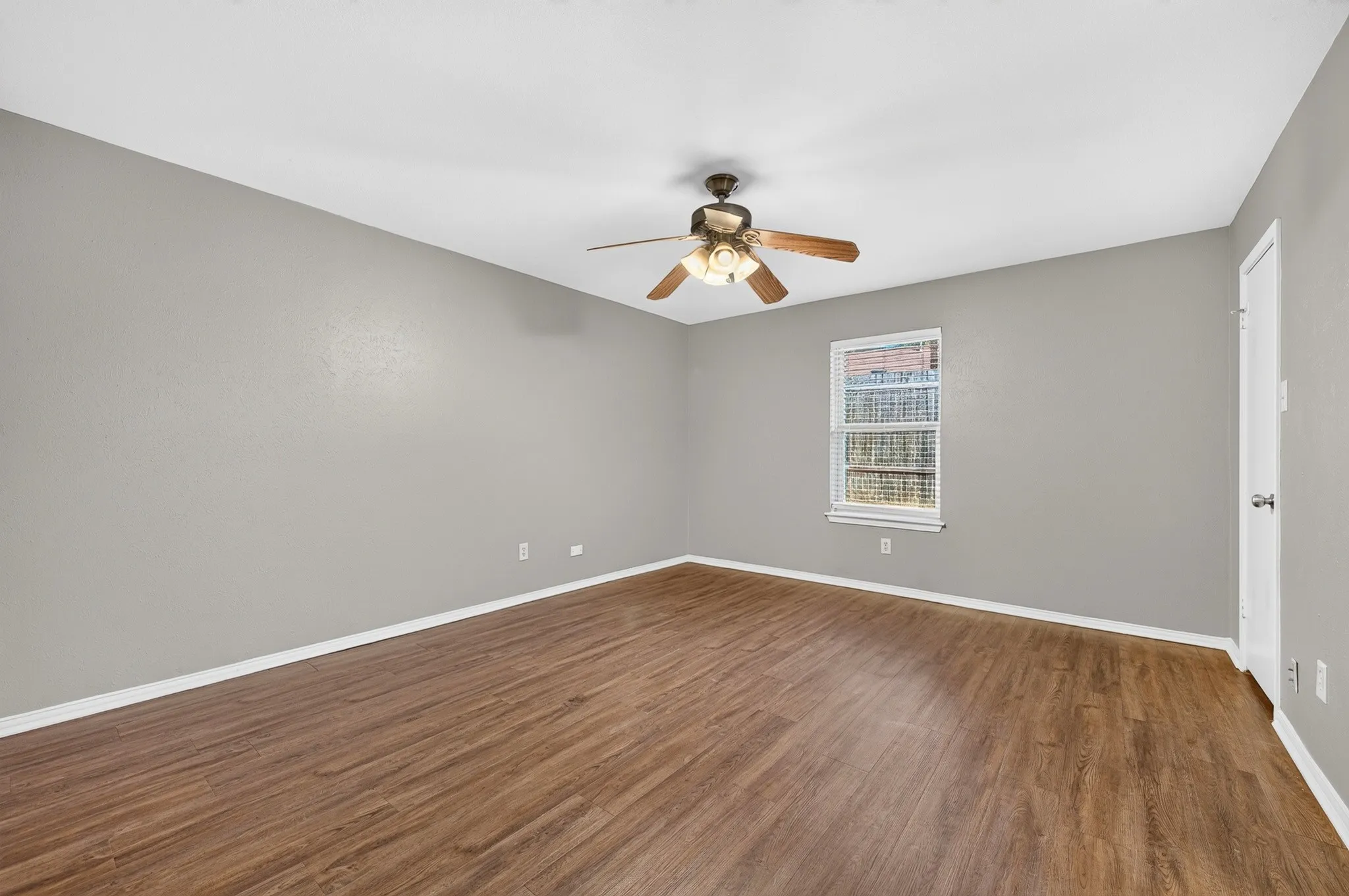 Spacious room featuring wood-finish flooring, light gray walls, a ceiling fan with integrated lighting, and a window for natural illumination
